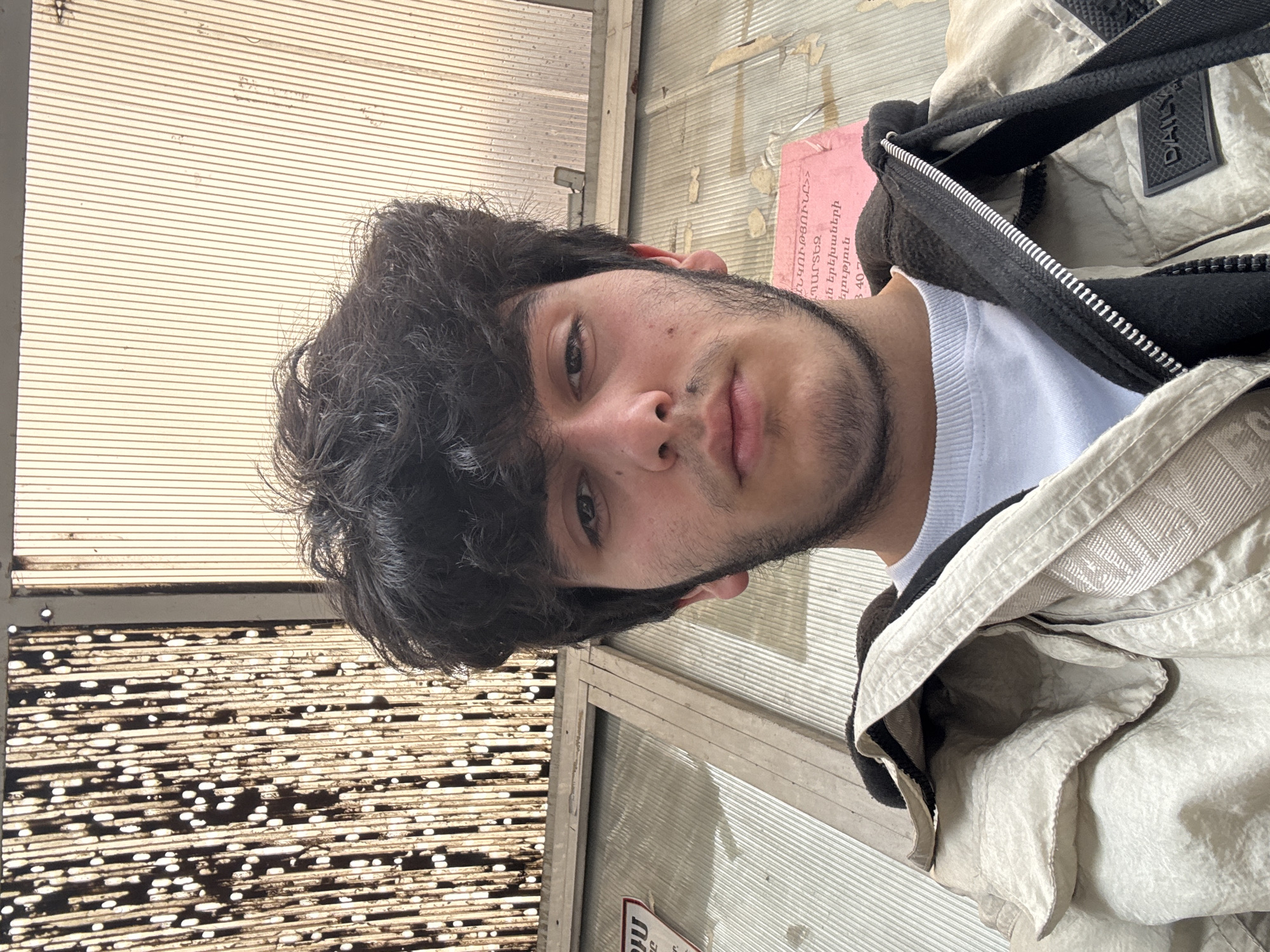 A young man stands in what appears to be a bus stop in Yerevan, Armenia. The bus stop has a metal frame and the back of the stop appears to be white metal slats. In the background are dirty metal bars with dots that are reflective and backlit by the sun. The man is wearing a white t-shirt, black sweatshirt, and a light beige jacket. He appears to be looking at the camera. A sign with Armenian text is visible on the structure of the bus stop.