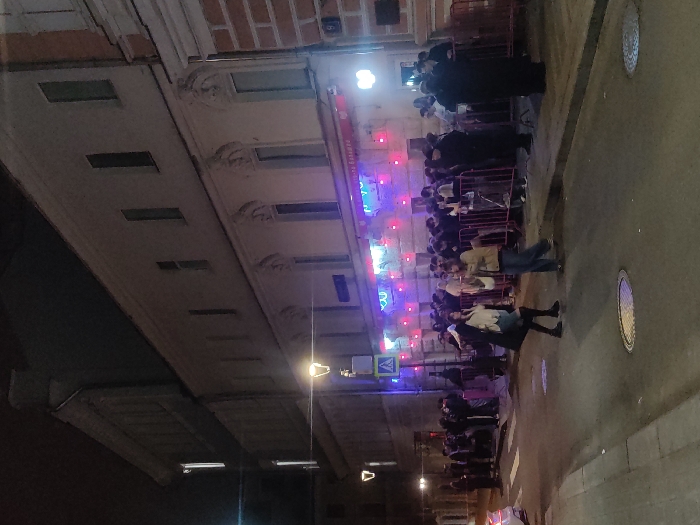 The image depicts a street scene in Moscow, Russia, at night. A queue of people stands outside a building illuminated with colorful neon lights. Pedestrians walk on the street beside the queue. The architecture consists of multi-story buildings with detailed facades. Streetlights cast a warm glow along the road. A pedestrian crossing sign is visible. The pavement has manhole covers.