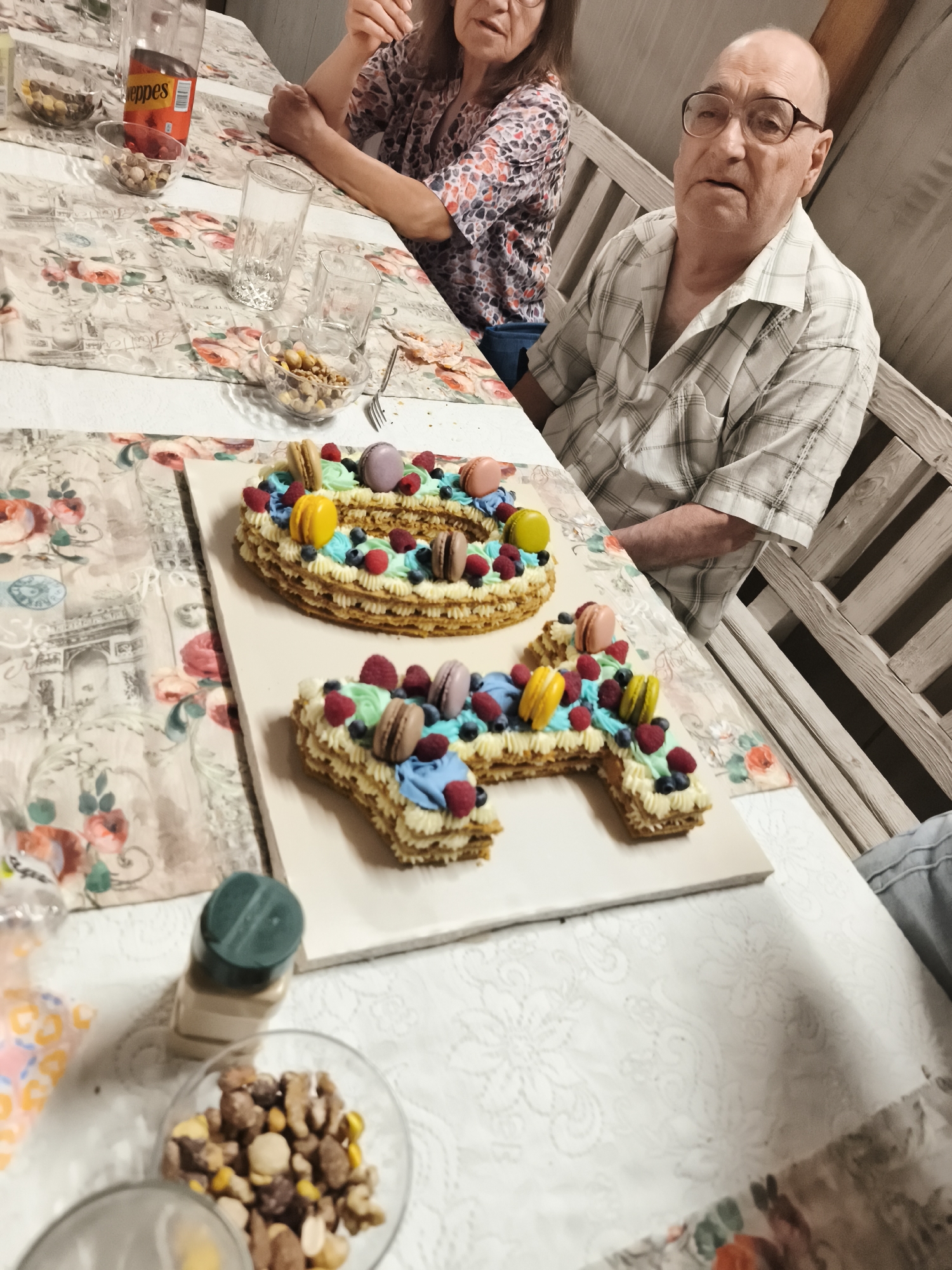 This media file captures a birthday celebration, likely for a milestone birthday, as indicated by the number cake which appears to be shaped into the numbers "6" and "4". Two people are partially visible: an older man with glasses and a woman in a floral shirt. The man is seated at a table, looking towards the camera with a neutral expression. The woman is seated behind him, her arms crossed.
The scene is set indoors at a table adorned with a floral tablecloth. On the table, in addition to the cake, are glasses, bowls of nuts, and a bottle of "Seppes" branded drink. The cake itself is elaborately decorated with cream frosting, fresh berries (raspberries and blueberries), and colorful macarons. The cake is presented on a white board.
The lighting suggests it could be daytime or early evening indoors. The overall atmosphere appears to be a family gathering or a casual celebration. The presence of a cake and celebratory food indicates a special occasion. The man's attire, a checkered shirt, and the wooden bench in the background suggest a comfortable, possibly rustic or home setting. The backdrop also hints at the location context provided: Stamboliyski, Bulgaria, possibly a home or a restaurant with a traditional or cozy ambiance.