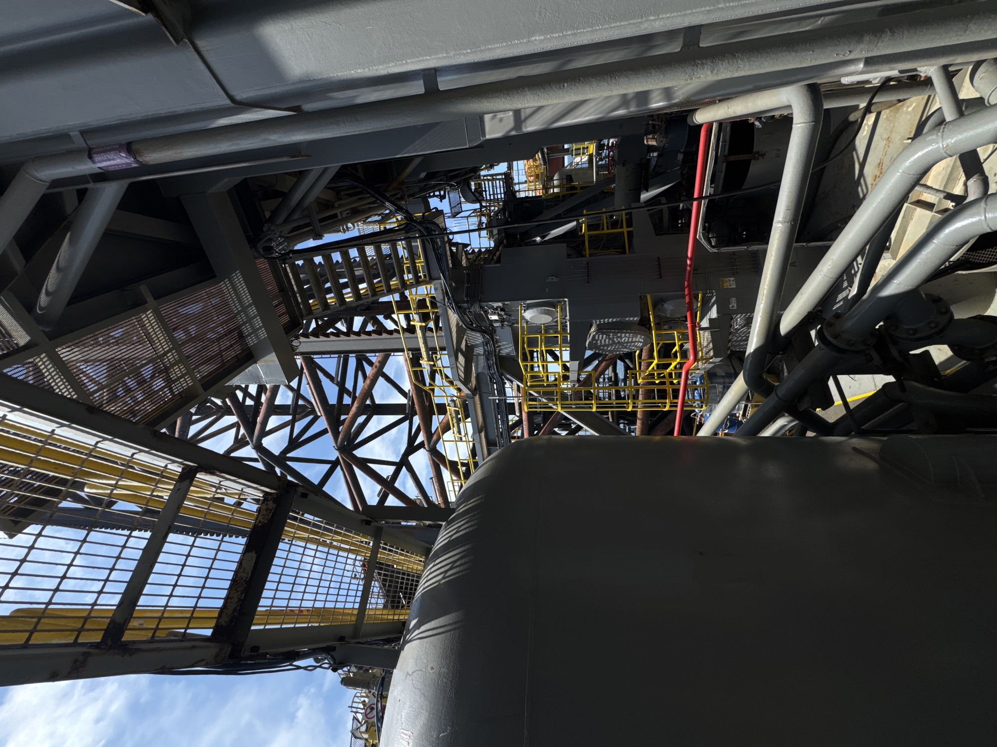 The image shows an upward-looking view of industrial infrastructure, possibly a section of an oil rig or a large processing plant. The scene is dominated by various metal structures, including large pipes, walkways with yellow railings, and supporting beams. The overall color palette consists of grays, yellows, and hints of red from some cables.
In the foreground, a large, curved, gray object, possibly a tank or a large pipe, takes up a significant portion of the lower right of the frame. Above this, a complex network of pipes and metal structures extends upwards and outwards. Yellow guardrails are visible on elevated platforms and walkways, suggesting a multi-level industrial environment. Some sections of the metalwork appear aged and possibly rusty, while other parts, like the pipes, are painted gray and appear to be in better condition.
The sky is visible through the gaps in the structures, indicating it is daytime. The sky is blue with some scattered white clouds, suggesting fair weather. The lighting suggests it is likely daytime, with sunlight casting shadows on the structures.
There is no visible activity or people in the image. The focus is entirely on the static industrial components.
A faint label with purple lettering is visible on a pipe in the upper left portion of the image, but the text is too blurry to read. Another small, partially obscured sign with red and white markings is visible on the lower right.
The image appears to be taken from ground level, looking straight up into the complex machinery of a large industrial facility. The perspective gives a sense of scale and the intricate nature of such structures.