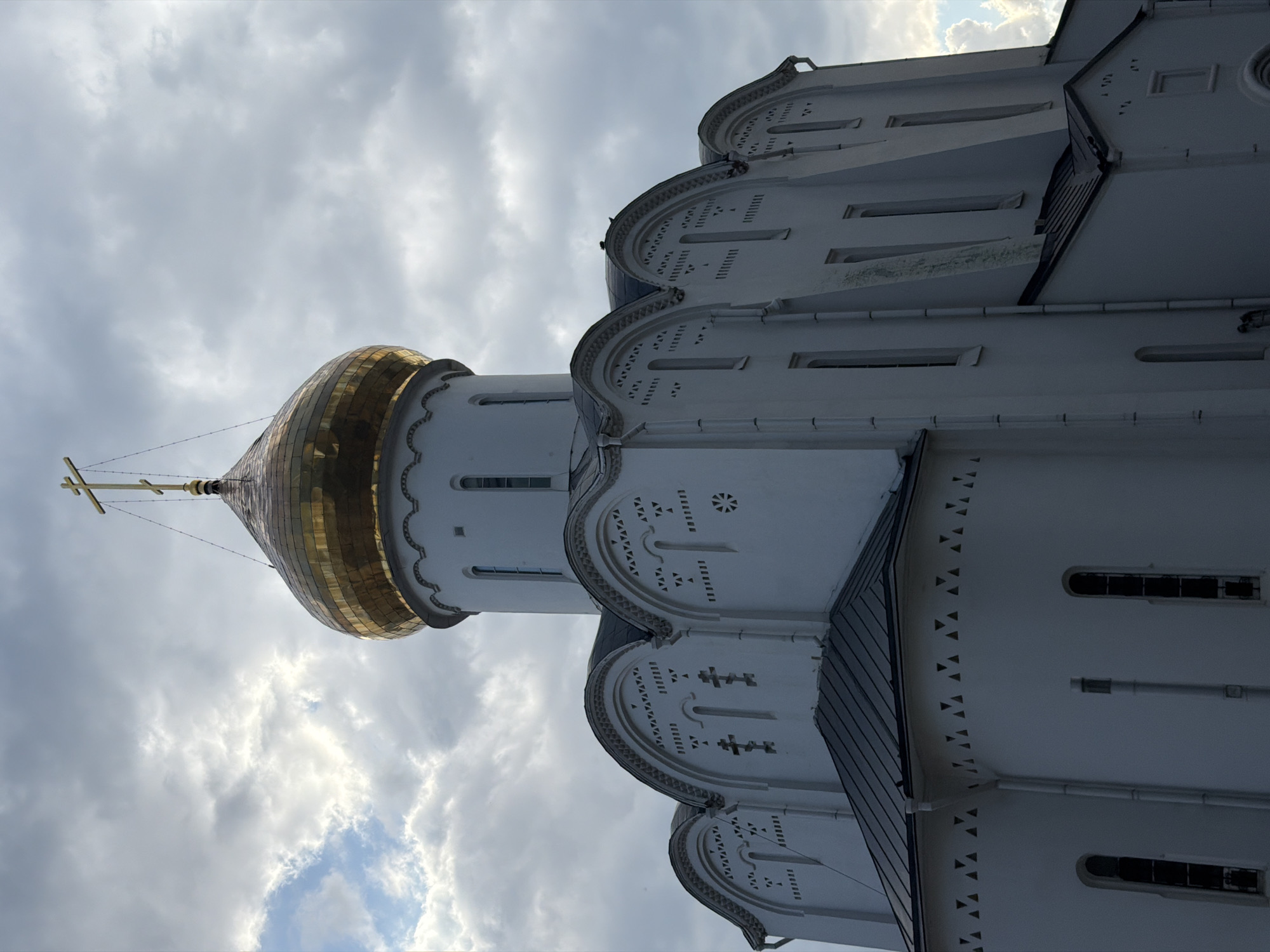 The image shows a white Orthodox church with a golden dome and a cross against a cloudy sky. The church has arched windows and decorative patterns. The architecture is traditional for Russian Orthodox churches. The specific location in Moscow is not identifiable from the image alone.