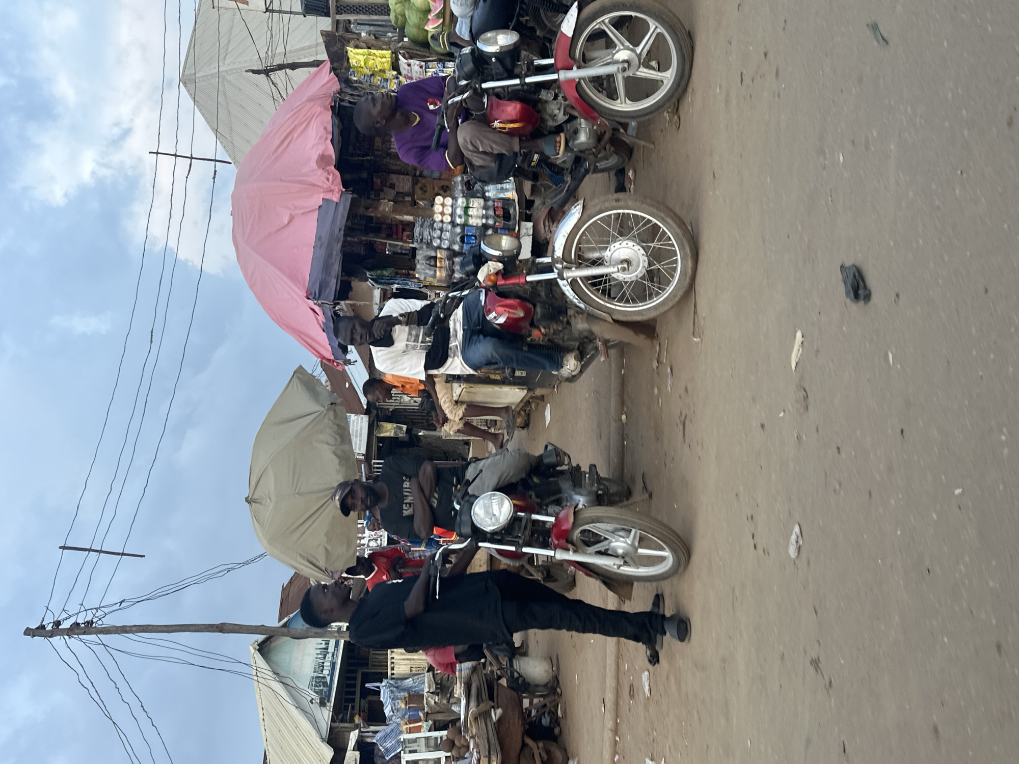 In a street market scene in Abuja, Nigeria, several men are gathered around motorcycles. In the foreground, a man in a black t-shirt and black pants stands beside a red motorcycle. To his right, a man in a black t-shirt with "KENUPS" written on it, wearing a baseball cap and grey pants, is seated on another red motorcycle. Further to the right, a man in a purple t-shirt and brown pants is also seated on a red motorcycle. Behind them, stalls are set up with goods, including bottles of drinks, and some fruits such as watermelons are visible. Two large umbrellas, one beige and one pink, provide shade over the stalls. Overhead, power lines crisscross the sky, supported by a wooden pole. The ground is dusty and unpaved. The sky is cloudy.
