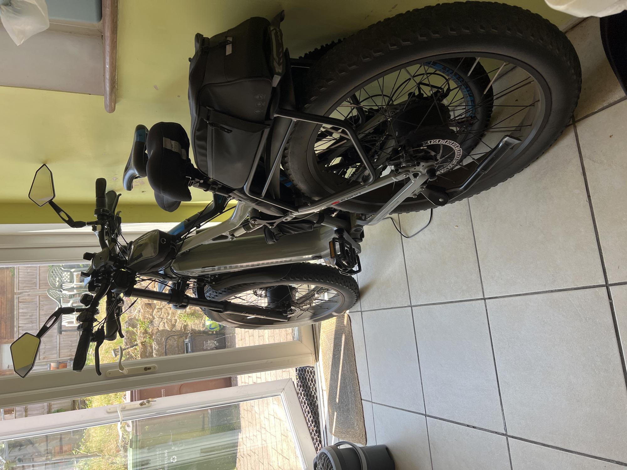 The image shows a light grey electric bicycle parked indoors, near a set of double glass doors that open to an outdoor patio area. The bicycle is positioned facing away from the doors, with its rear wheel closest to the camera. It has large, knobby tires, a rear rack with a black storage bag, and a second seat behind the main seat. The handlebars are visible, with mirrors attached. The interior features pale tiled flooring and a pale yellow wall. The outdoor area beyond the doors appears to be a residential garden with some outdoor furniture. The location is identified as being in Guildford, United Kingdom. There are no people visible in the image.