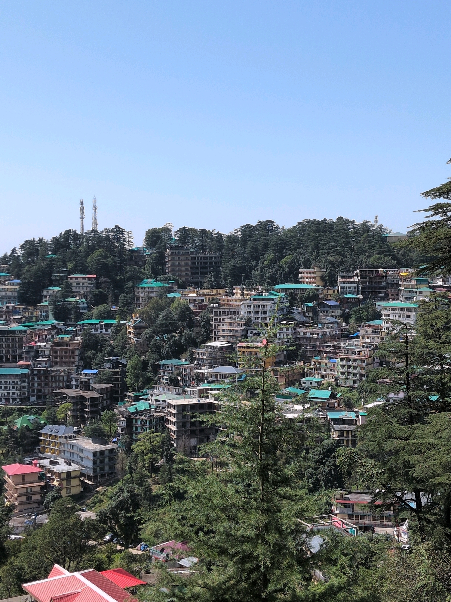 The image depicts a dense hillside town, likely Bhagsu, India, characterized by numerous buildings and lush green trees. The architecture is varied, with multi-story buildings featuring flat roofs and some with pitched, often green-colored, roofs. Many structures appear to be under construction or in varying states of completion, with exposed concrete and unfinished facades visible. The town is built on a steep incline, with buildings cascading down the hillside. Evergreen trees are interspersed among the buildings, and in the foreground, large coniferous trees partially obscure the view. Two prominent telecommunication towers are visible on a distant ridge, against a clear blue sky. The scene suggests a vibrant, perhaps rapidly developing, urban environment nestled within a mountainous, forested landscape. There are no people or animals clearly visible in the image.