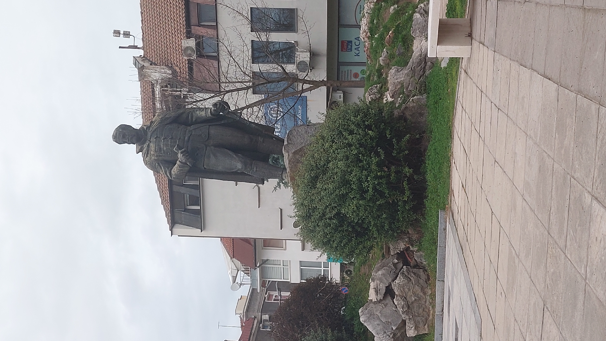 The image depicts a bronze statue of a man standing in a plaza in Gabrovo, Bulgaria. The statue is of a figure in a military-style uniform with a buttoned coat and appears to be holding something in his left hand. It stands on a stone base among landscaping that includes bushes, grass, and rocks. Behind the statue is a building with a red tile roof and white walls. Air conditioning units are visible on the building's exterior. A sign is present, but the text is difficult to read. The foreground consists of a paved area with light-colored rectangular stones. The sky is overcast.