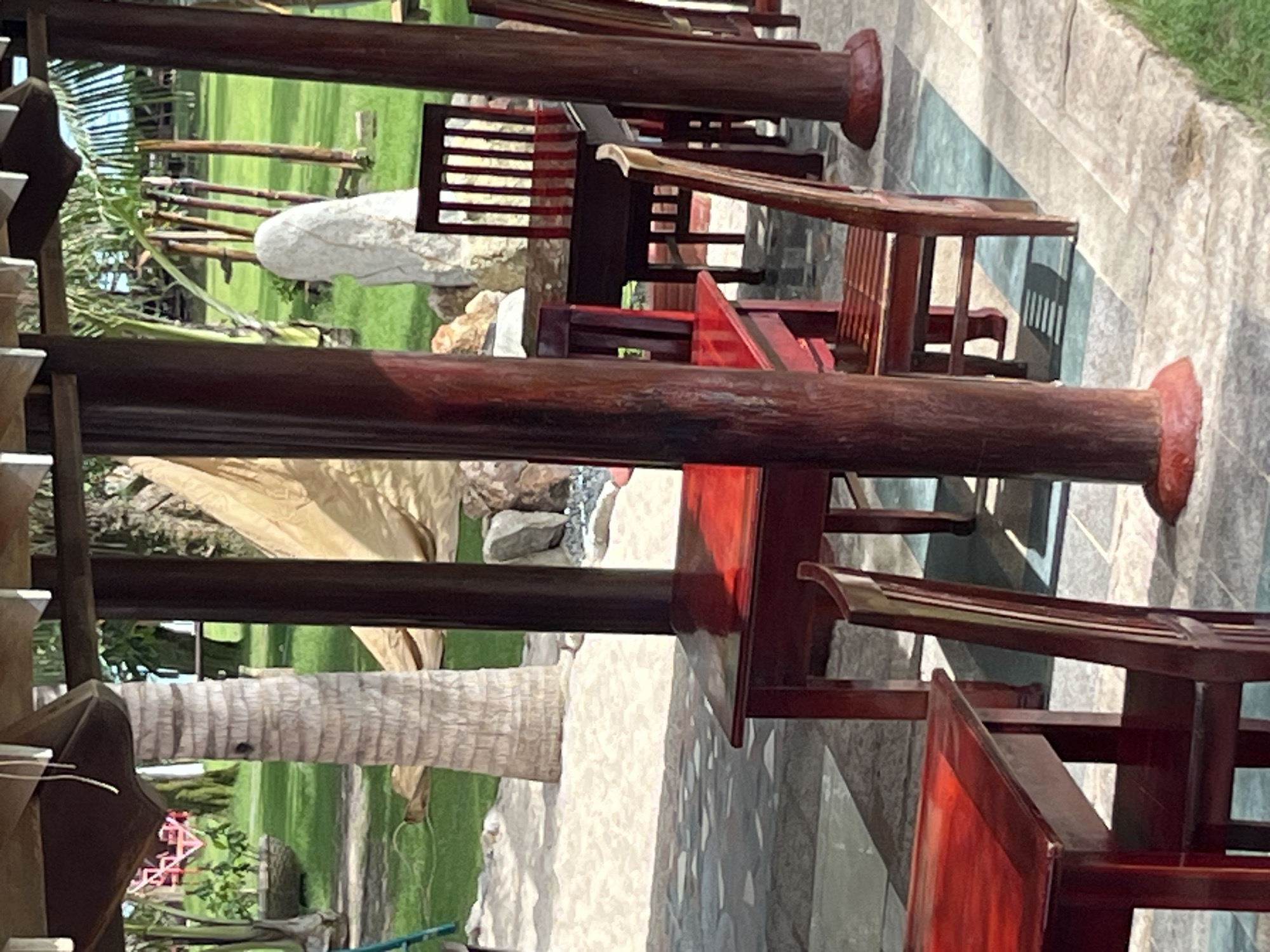 This image depicts an outdoor seating area at what appears to be a resort or restaurant in Mũi Né, Vietnam. The scene is characterized by several wooden tables and chairs, primarily in a dark red or brown finish. The tables are set on a paved ground with geometric patterns. In the foreground, a red table with a chair is visible. Further back, another red table with a chair is positioned. A dark brown table with matching chairs is seen in the mid-ground, set on a patterned stone floor.
The background reveals a lush green lawn with palm trees and other vegetation, typical of a tropical coastal environment. A large, light-colored stone sculpture stands prominently on the lawn. In the distance, what seems to be a sandy area can be observed, suggesting proximity to a beach. The overall ambiance is one of a tranquil outdoor dining or relaxation space, likely designed to complement the natural beauty of Mũi Né. No people are present in the image.