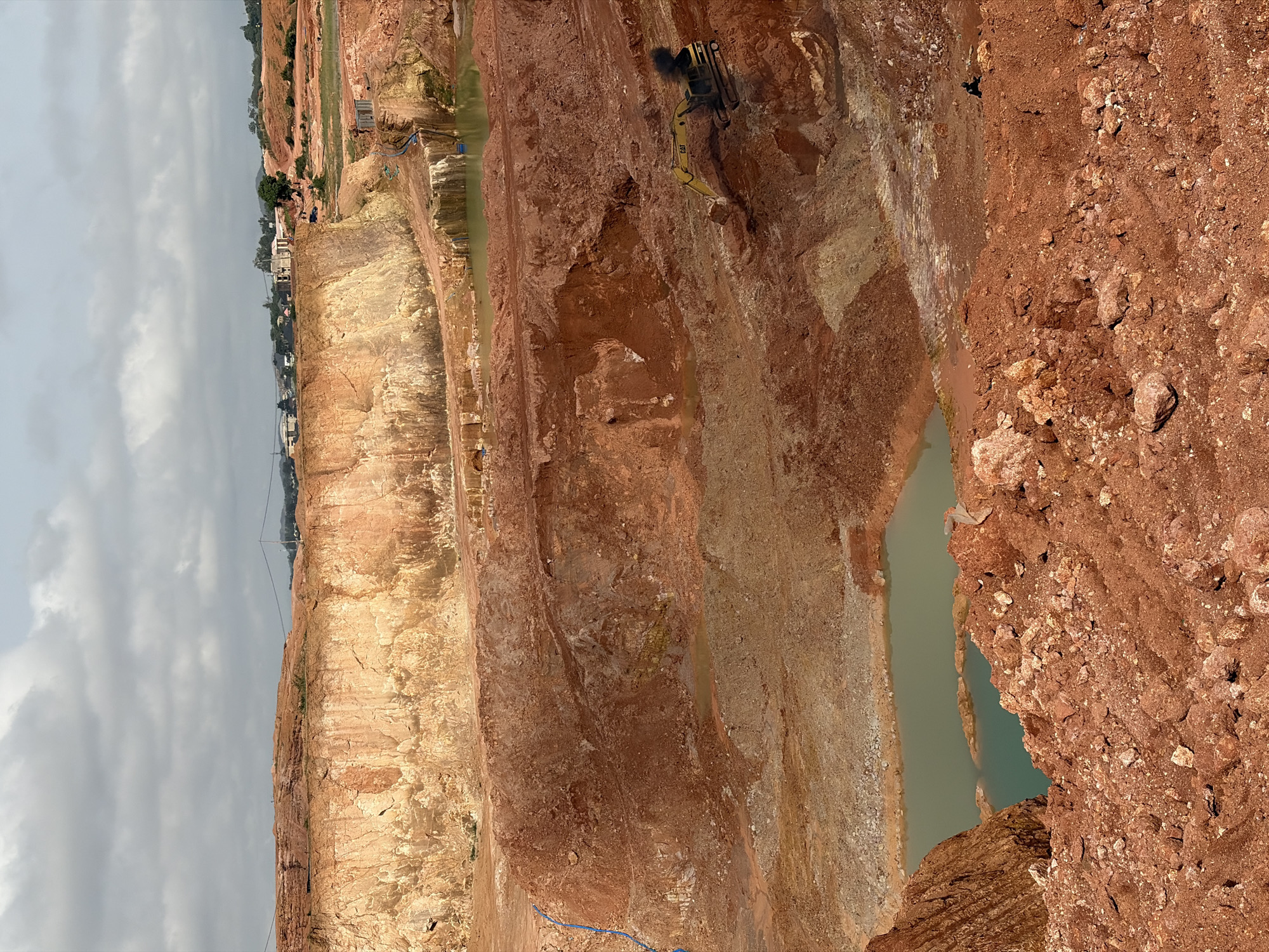 This is a medium-shot, high-angle, full-color photograph of an active quarry in Kwang, Nigeria, under a cloudy sky. The quarry features massive, terraced excavations of reddish-brown earth, with lighter, yellowish-beige rock formations visible at the upper levels. Several pools of murky, greenish water have accumulated at different depths within the quarry.
In the middle ground, a yellow excavator with a black plume of smoke emanating from its exhaust is positioned on a lower tier of the quarry, its boom extended. Further back, a small number of figures, appearing as dark silhouettes, are visible on the upper rim of the quarry and near some structures. Some of these structures appear to be simple buildings under construction or storage units. A blue pipe is visible running down the side of one of the excavated walls. In the distance, a settlement with scattered buildings and trees can be seen on a low-lying hill under the overcast sky. The foreground is dominated by the rough, reddish-brown soil and rocks of the quarry's edge.
