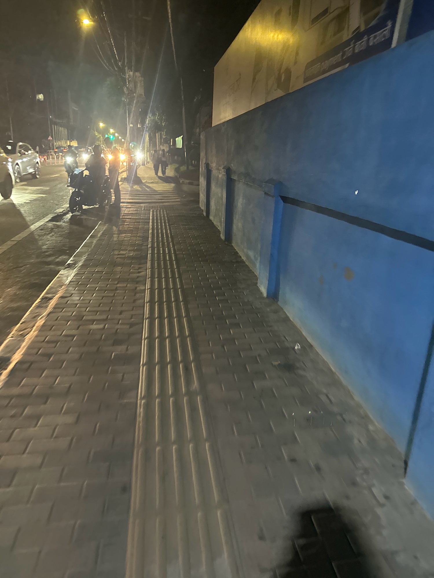 The image depicts a street scene at night in Kathmandu Metropolitan City, Nepal. A paved sidewalk with tactile paving for the visually impaired runs along the right side of the frame, adjacent to a bright blue wall. The sidewalk is illuminated by streetlights, casting long shadows. Across the street, there is traffic, including cars and motorcycles, with their headlights and taillights visible. Several individuals are present on the sidewalk and near the vehicles. One group appears to be conversing near a parked motorcycle, while others walk along the sidewalk. A large, illuminated sign with text in a non-Latin script is mounted on the wall in the background. The overall impression is of a busy urban environment at night.