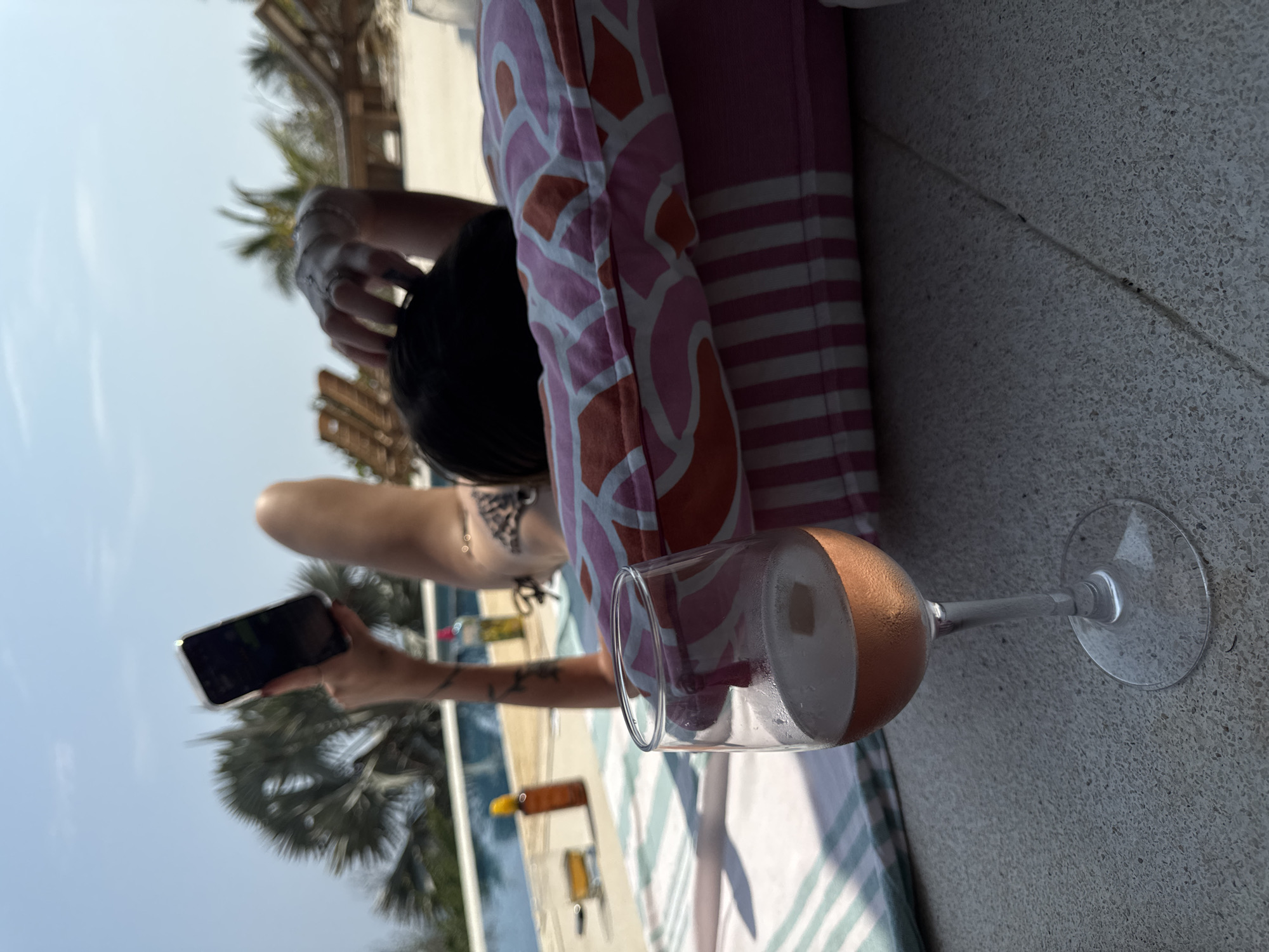 A woman is lounging by a pool in Unknown, Mexico, propped up against a pink pillow. She is holding a cell phone in her left hand, presumably taking a picture. Beside her is a wine glass filled with a pink drink and ice. In the background, there are palm trees and a glimpse of other buildings. The scene appears to be outdoors on a sunny day.