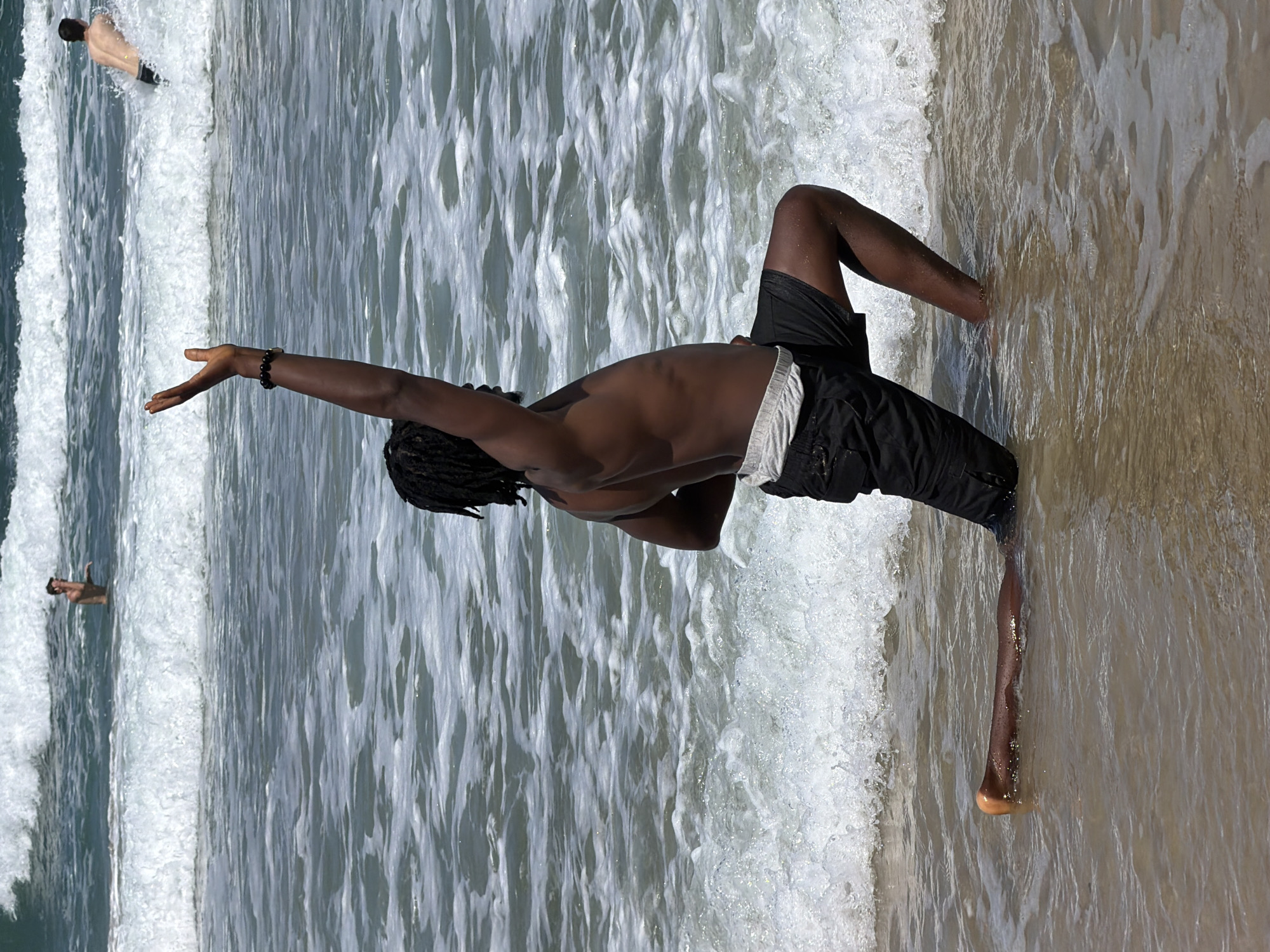 The image captures a scene on a beach in Tel Aviv, Israel. A dark-skinned individual, wearing black shorts and a white waistband, stands at the water's edge with one arm extended towards the sea. The person has dreadlocks. Further out in the ocean, two other people are visible swimming in the waves. The beach appears to be sandy, and the water shows signs of waves and foam. The image orientation is rotated.