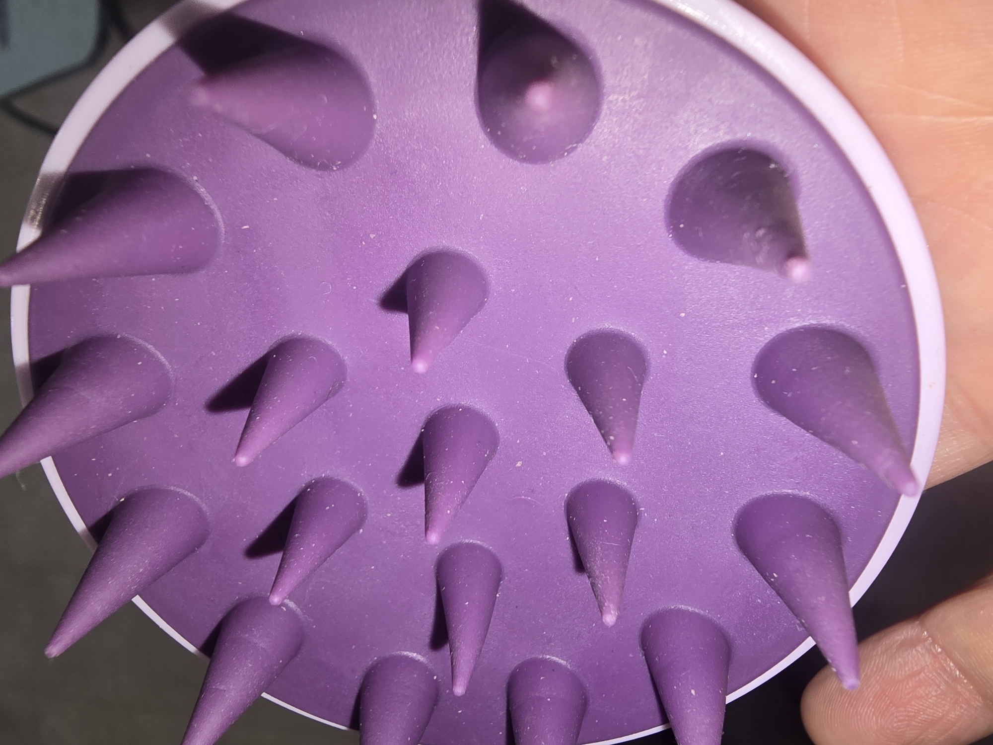 A close-up, overhead shot shows a purple silicone scalp massager held by a person's hand. The massager has numerous conical nubs. The background is dark and out of focus. The scene is not in Ulan-Ude, Russia.