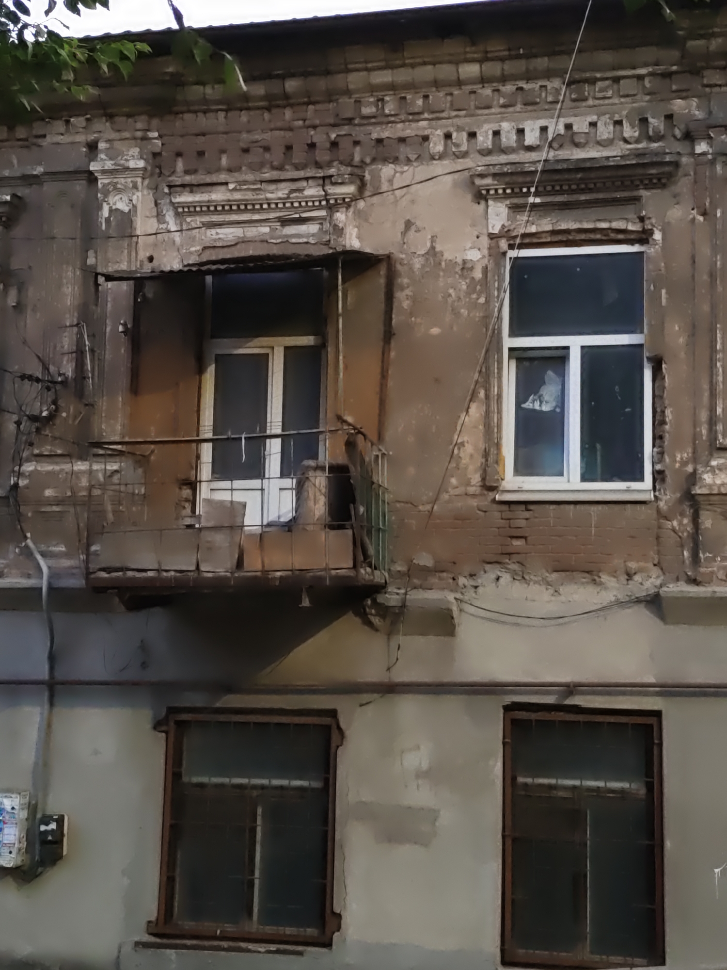 The image shows the facade of an old, dilapidated apartment building in Rostov-on-Don, Russia. The building's brickwork is exposed in places, with peeling paint and visible signs of decay. There is a balcony on the second floor with a rusty metal railing, partially covered by what appears to be cardboard boxes. A white door is visible on the balcony. To the right of the balcony is a window with a white frame. Below the balcony and the window, on the first floor, are two windows with metal bars, suggesting security measures or perhaps simply old, reinforced frames. The windows on the first floor are dark, their interiors obscured. An electrical meter or fuse box is mounted on the wall to the left of the first-floor windows. Green leaves from a tree hang into the frame from the top left corner. The overall impression is one of neglect and a bygone era.