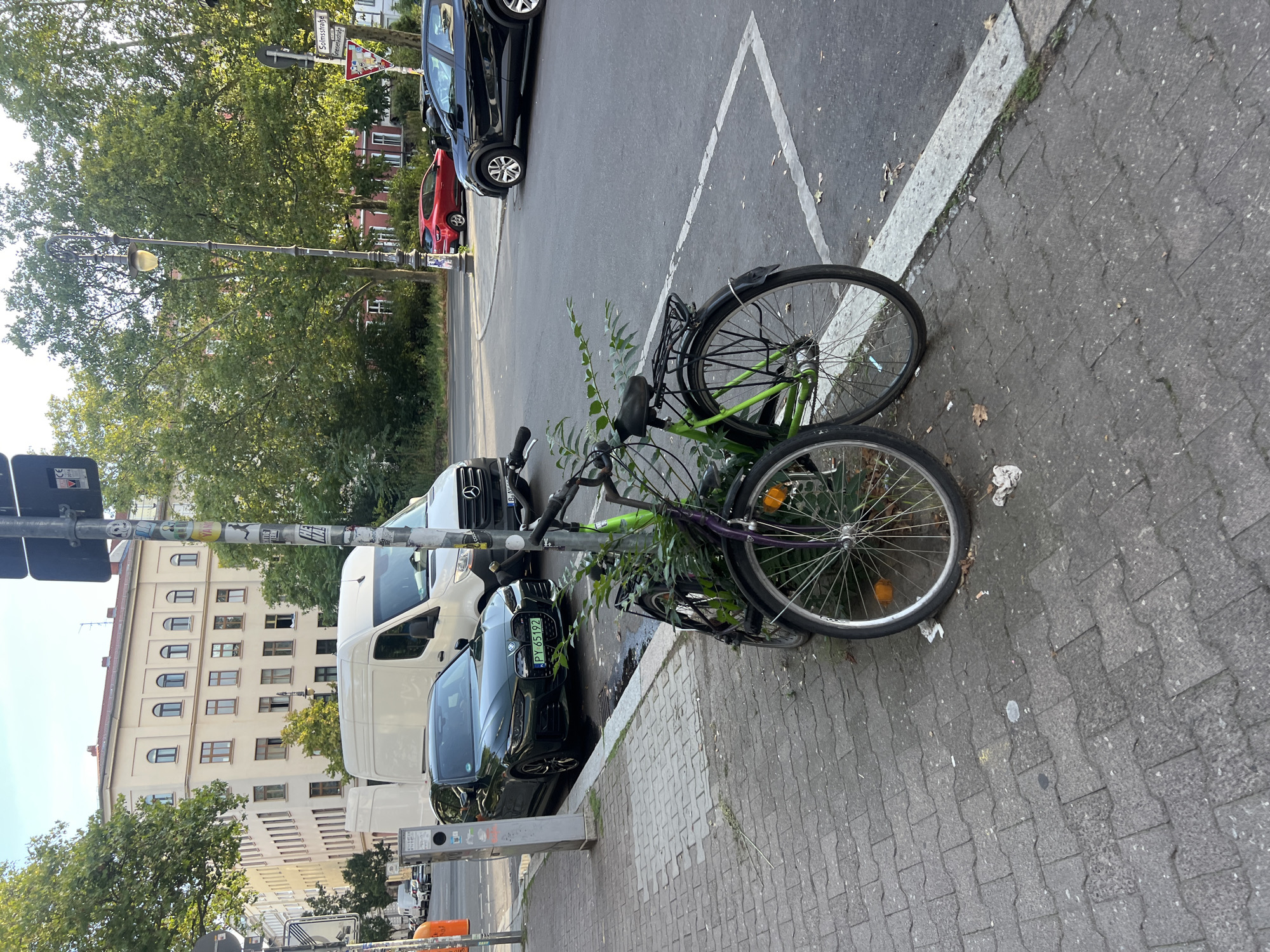 The image shows a street scene in Berlin, Germany. In the foreground, two parked bicycles are leaning against a pole. One bicycle is green and the other is purple. Behind the bicycles, a dark green BMW car is parked on the side of the road, with a license plate that reads "PY 65192". Further back, a white van is parked. In the background, there are buildings and trees. The sky is clear and blue. There are no people visible in the image. The scene appears to be a typical urban street in a residential area. The street is paved with cobblestones and the curb is made of concrete. There are traffic signs and streetlights visible. The overall impression is one of a quiet, ordinary day in the city.