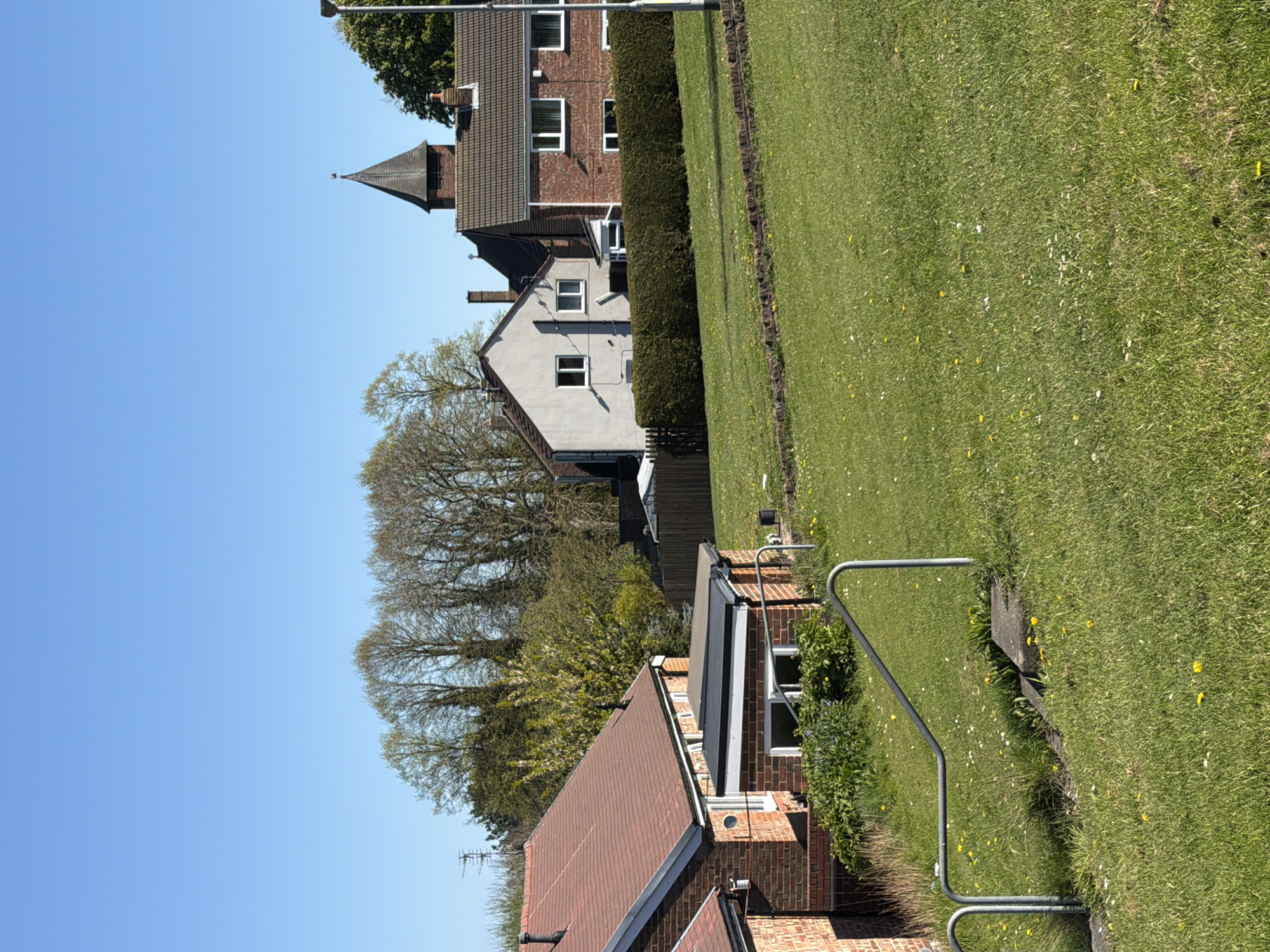 This media file captures a sunny, clear day in a residential area of Bearpark, United Kingdom. The scene features a row of houses bordered by a wide, green grassy area.
On the left side of the image, several houses are visible. One prominent house in the mid-ground features red brick walls and a distinctive dark, pointed, or conical roof structure, resembling a small spire or turret. Below this, another house has light-colored walls and a dark roof. In the foreground, partially visible, is a house with a reddish-brown tiled roof and brickwork, displaying a television aerial. Between the houses and the grassy expanse runs a neatly trimmed green hedge.
The right side of the image is dominated by a large, well-maintained lawn, dotted with small white and yellow wildflowers, likely daisies and dandelions. A metal railing, possibly indicating a path or boundary, is present in the lower right foreground next to the grass. In the background, numerous deciduous trees, mostly bare or with very early foliage, rise against a bright, cloudless blue sky, which fills the upper portion of the image. A street light is visible near the top right.
The overall impression is one of a quiet and peaceful suburban or village setting under bright daylight. No human activity or visible text is present.