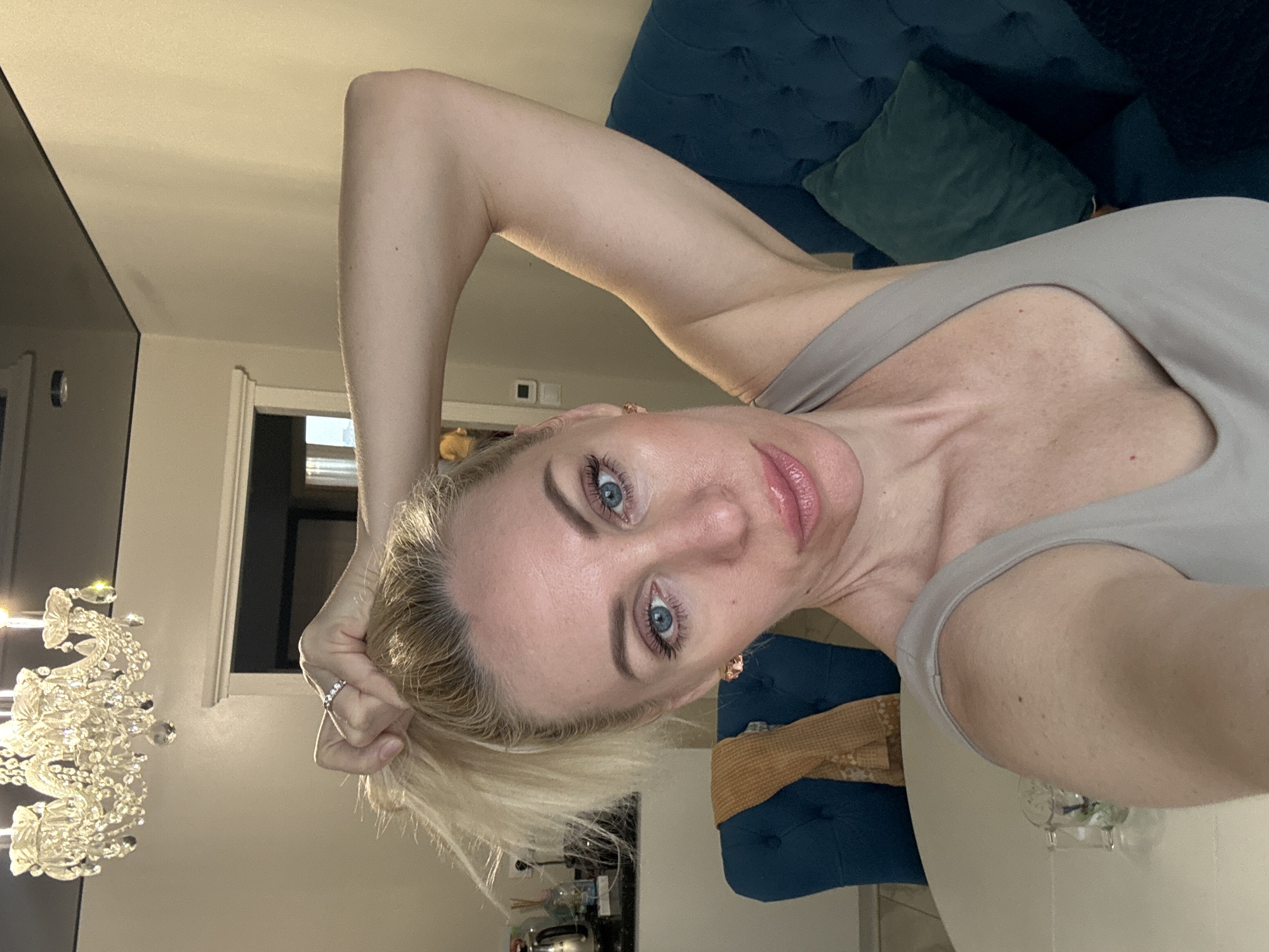 A fair-skinned woman with blonde hair and blue eyes is posing for a selfie. She is wearing a light grey top and a ring on her finger. Her right arm is raised above her head, pulling at her hair. A tufted blue sofa with a green pillow is visible in the background, along with a white table. A chandelier is partially visible in the lower left corner. The location is Unknown, Unknown.