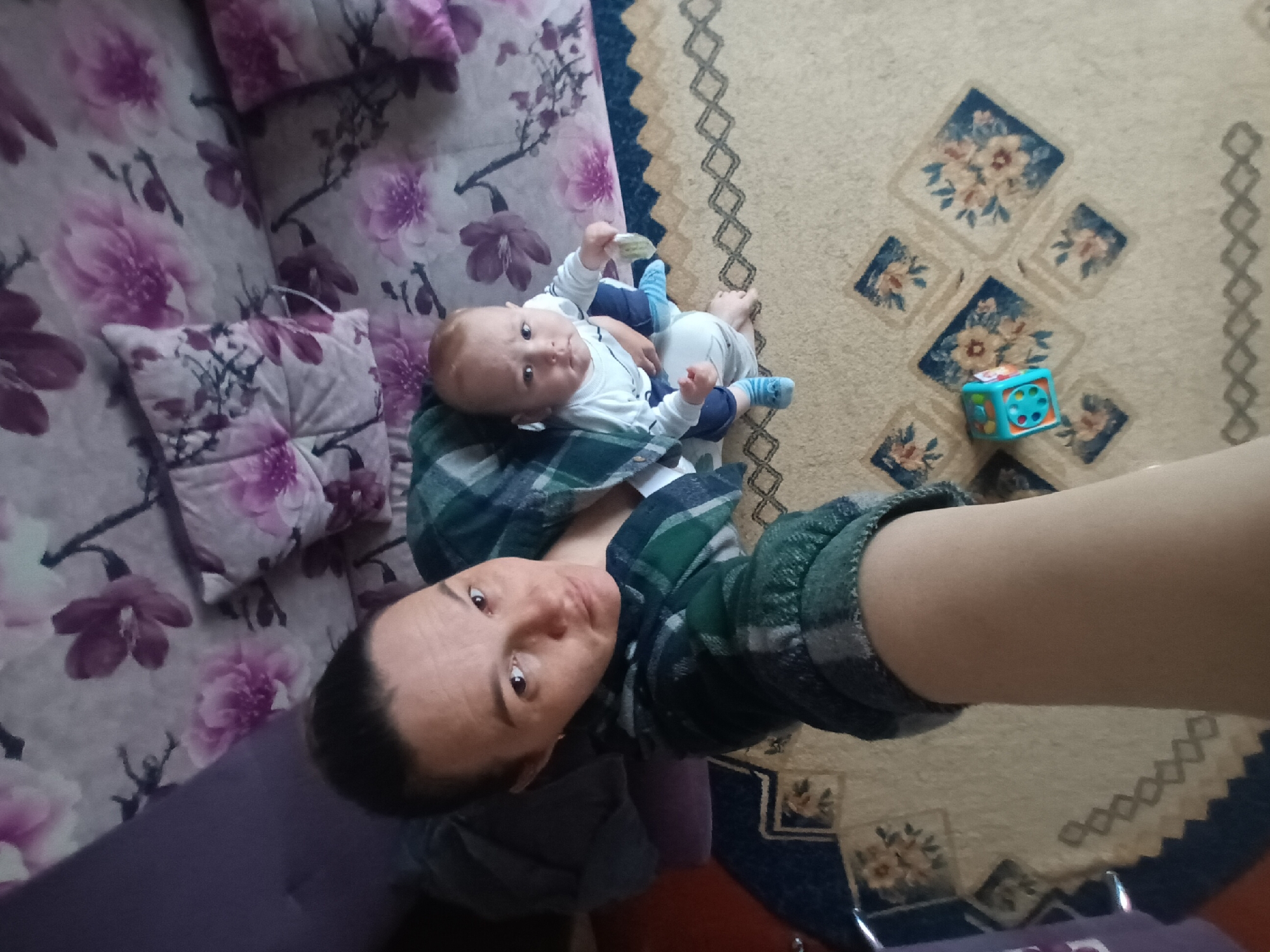 A selfie taken from a high angle in a residential space, likely in Ольховка, Russia, shows a light-skinned person in a green and gray plaid shirt holding a baby in a white and blue outfit. The baby is lying on the person's chest, looking towards the camera. To the right, there's a colorful toy cube on a beige carpet with floral patterns. A floral-patterned sofa is visible on the left.
