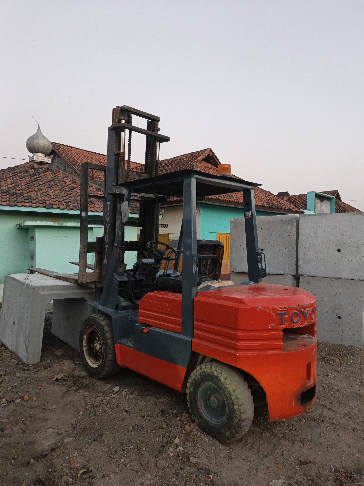 A red and gray Toyota forklift is parked on a dirt surface in front of a row of buildings with red tiled roofs. The forklift's forks are extended and resting on a large, rectangular concrete block. To the right of the forklift are stacks of similar concrete blocks. The buildings in the background appear to be residential or commercial structures, with varying colored walls and windows. The sky is overcast, suggesting it may be late afternoon or early morning. The scene is likely in Planggu, Indonesia, given the context of the prompt.