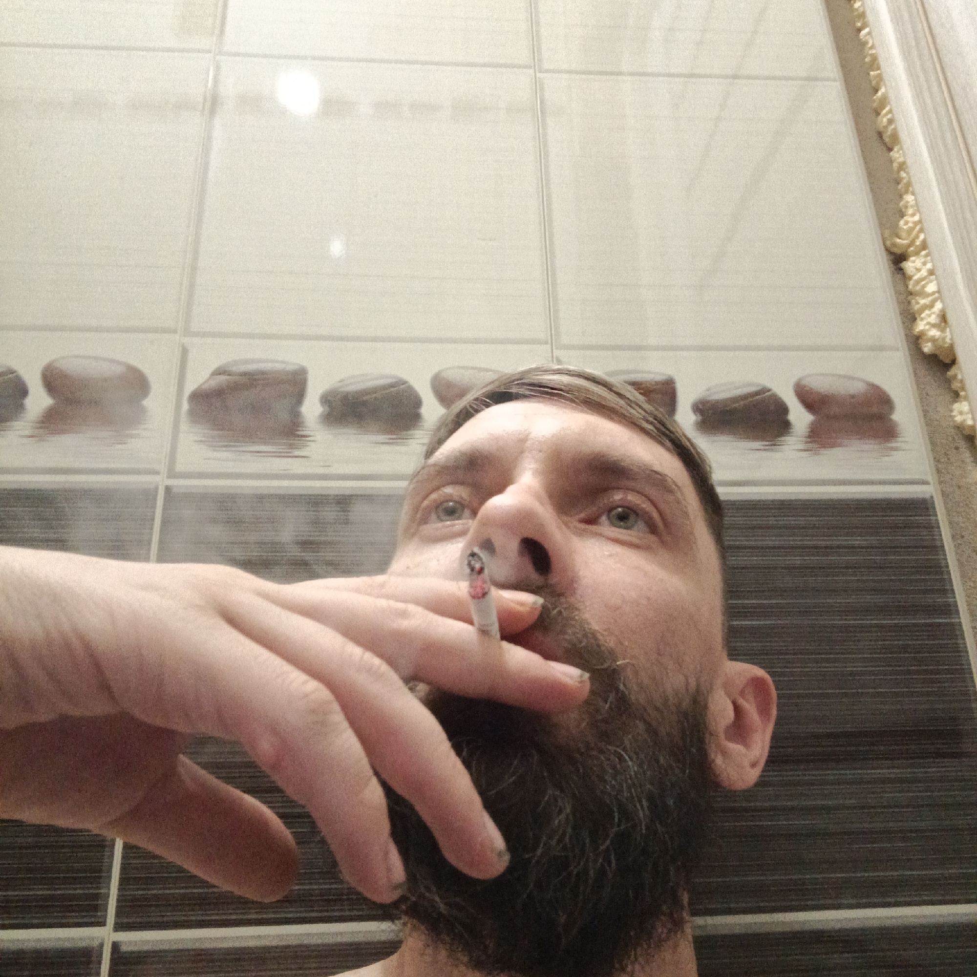 A man with a beard is holding a lit cigarette in front of his face, positioned against a background of bathroom tiles with a stone pattern. The location appears to be indoors. Марковский, Russia is not visually discernible in the image.