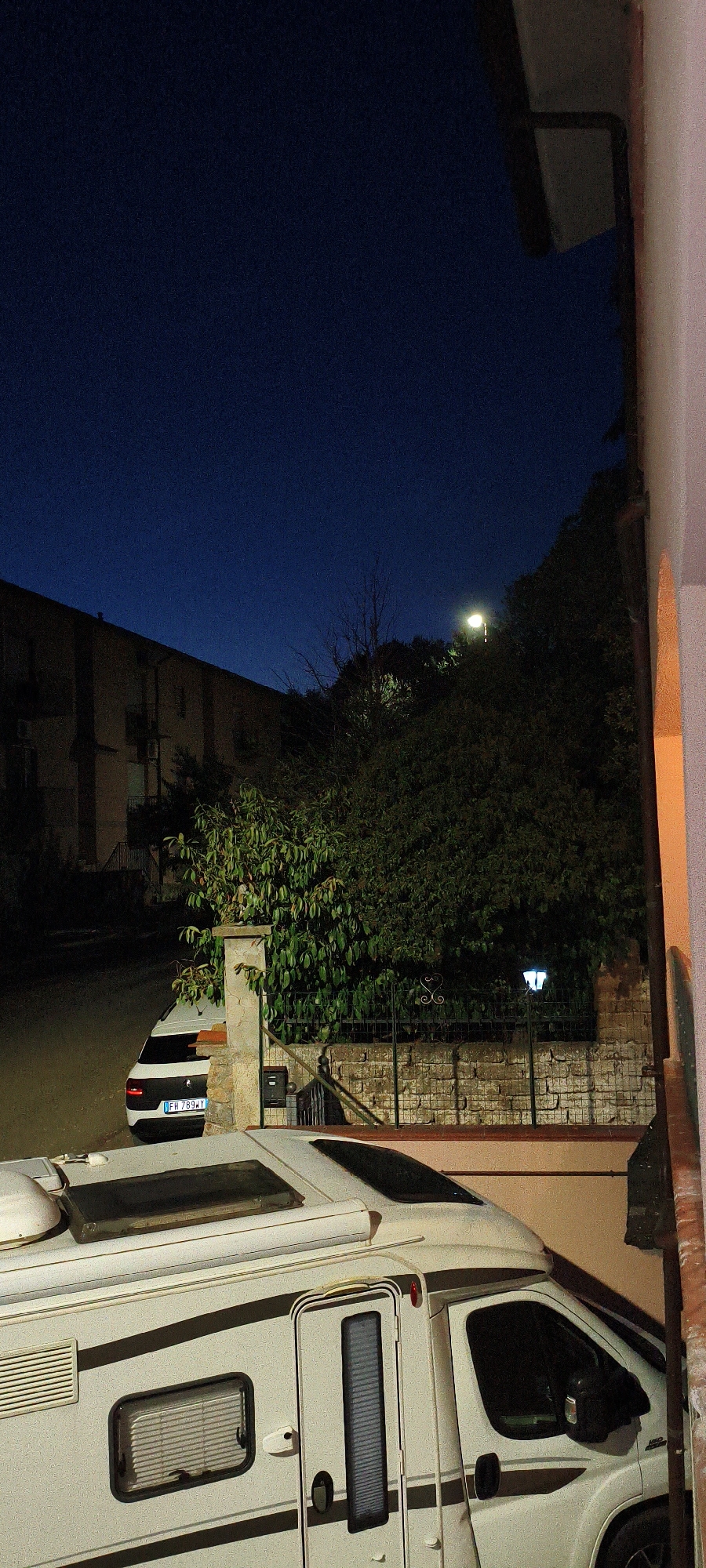 A white camper van is parked on a paved surface in front of a stone wall and a fence, with a white car parked behind it. The scene is illuminated by a street lamp and possibly the light from a building in the background. A dark blue sky with some faint light suggests it is dusk or dawn. The location appears to be a residential area with a building in the background. The camper van has several windows and a door, and the car has its license plate visible. The environment suggests a quiet, possibly rural or suburban setting in Campagnatico, Italy.