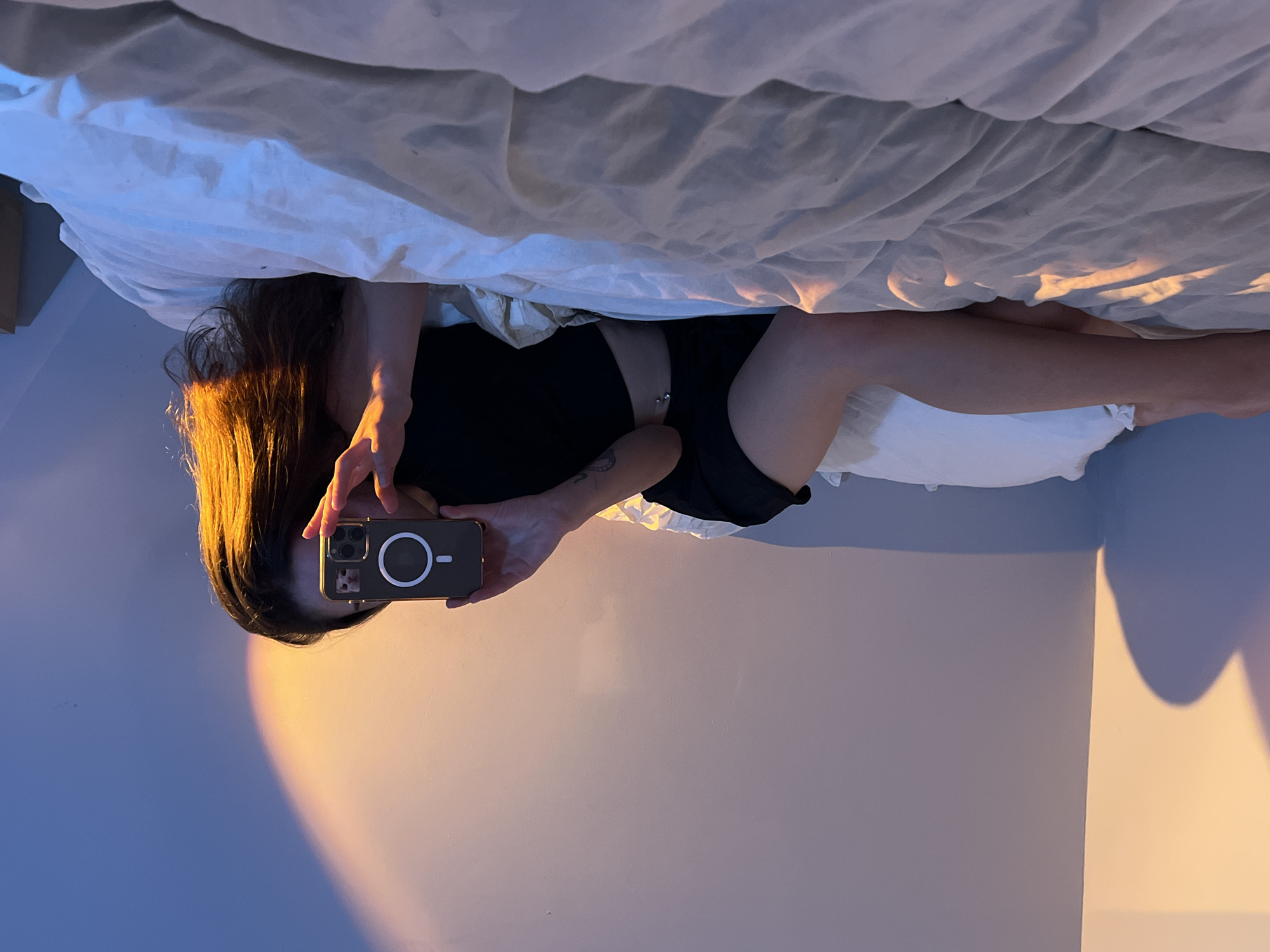 A person is lying on a bed, holding a smartphone in their hands to take a selfie. The person is wearing dark clothing and is illuminated by golden light. The room appears to be a bedroom with a light-colored wall and bedding. There are no specific details in the image that indicate a location in Ternopil, Ukraine.
