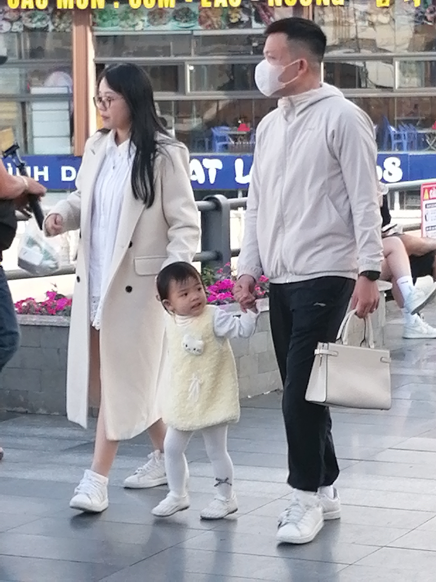 This image captures a family scene on a paved walkway in Da Lat, Vietnam. A woman wearing glasses, a white collared shirt, and a cream-colored trench coat walks on the left, holding a white plastic bag in her left hand. To her right, a young child in a light yellow sleeveless dress and white tights walks forward, looking to the right. A man in a light gray jacket, dark pants, and a white face mask walks on the right, holding the child's hand with his left hand and carrying a white handbag in his right hand. The background shows a building with glass windows and blue tables and chairs visible inside, suggesting a cafe or restaurant. Pink flowers are planted in a planter box to the left of the walkway. The ground is a gray tiled surface.
