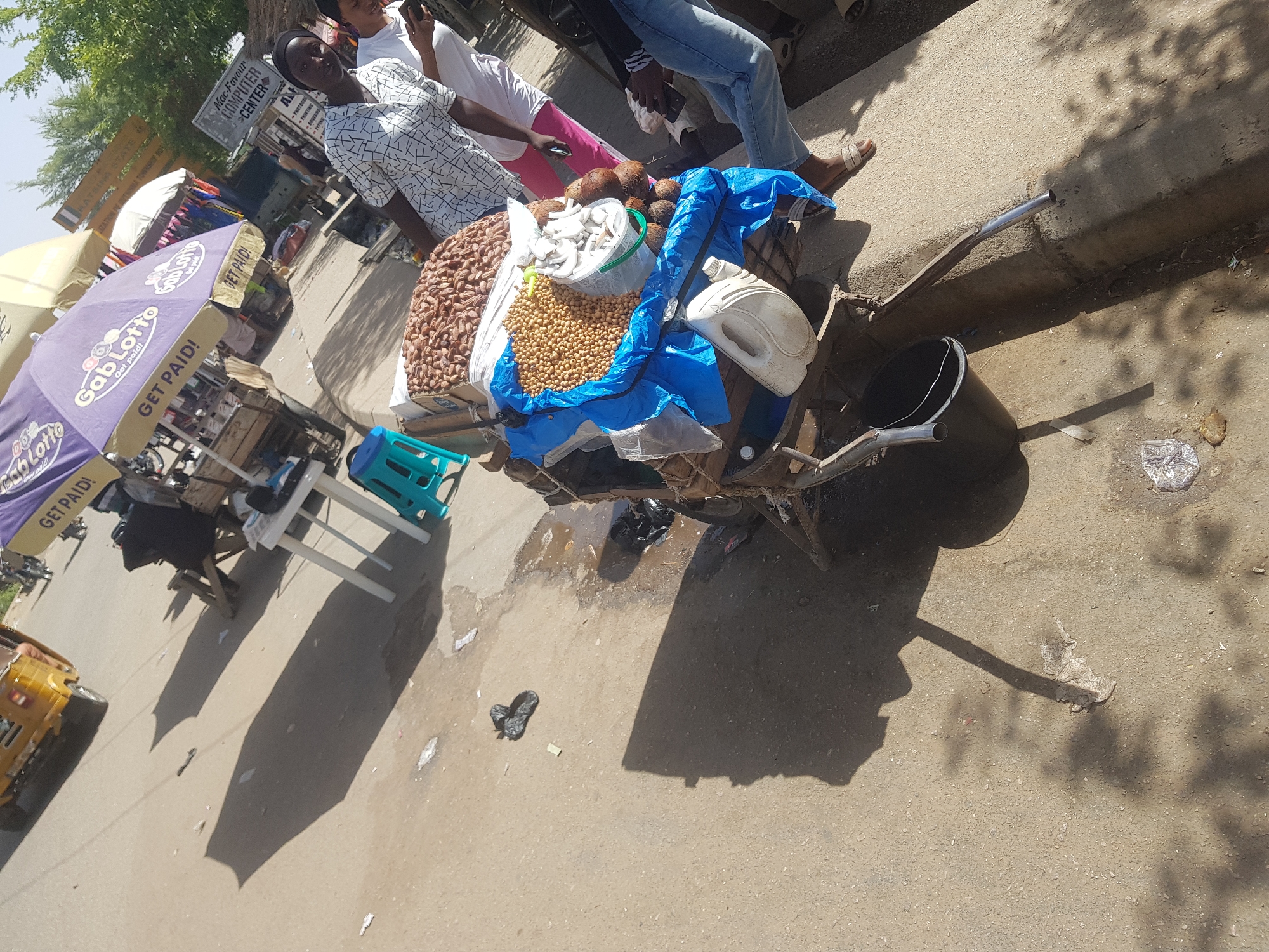 Here's a detailed description of the image:
**Location:** The image appears to be taken in Dabawa, Nigeria, based on sign reading "DABAWA STATE" and another mentioning "TOWNSHIP ROAD."
**People:**
* A man in a patterned shirt stands next to a hand-pushed cart.
* Other people are visible behind him, partially obscured. One is wearing a white shirt and pink pants.
* A person is wearing blue jeans and sandals.
**Objects:**
* Hand-pushed cart laden with goods. The cart holds peanuts, beans, coconuts (some with the flesh cut and in a bucket), and a white container. A blue tarp covers part of the merchandise.
* A black bucket hangs from the cart.
* A "Gab Lotto" umbrella is partially visible.
* A sign reads "Mac-Favour Computer Center" advertising services like photocopying, printing, and typing.
* A blue plastic stool and white plastic tables are present.
* A yellow tricycle vehicle is partially visible on the side of the road.
* Various pieces of litter are scattered on the ground.
**Environment:**
* The setting is an outdoor market area, likely near a road.
* The area appears to be a mix of concrete and dirt.
* Shadows indicate it is daytime.
