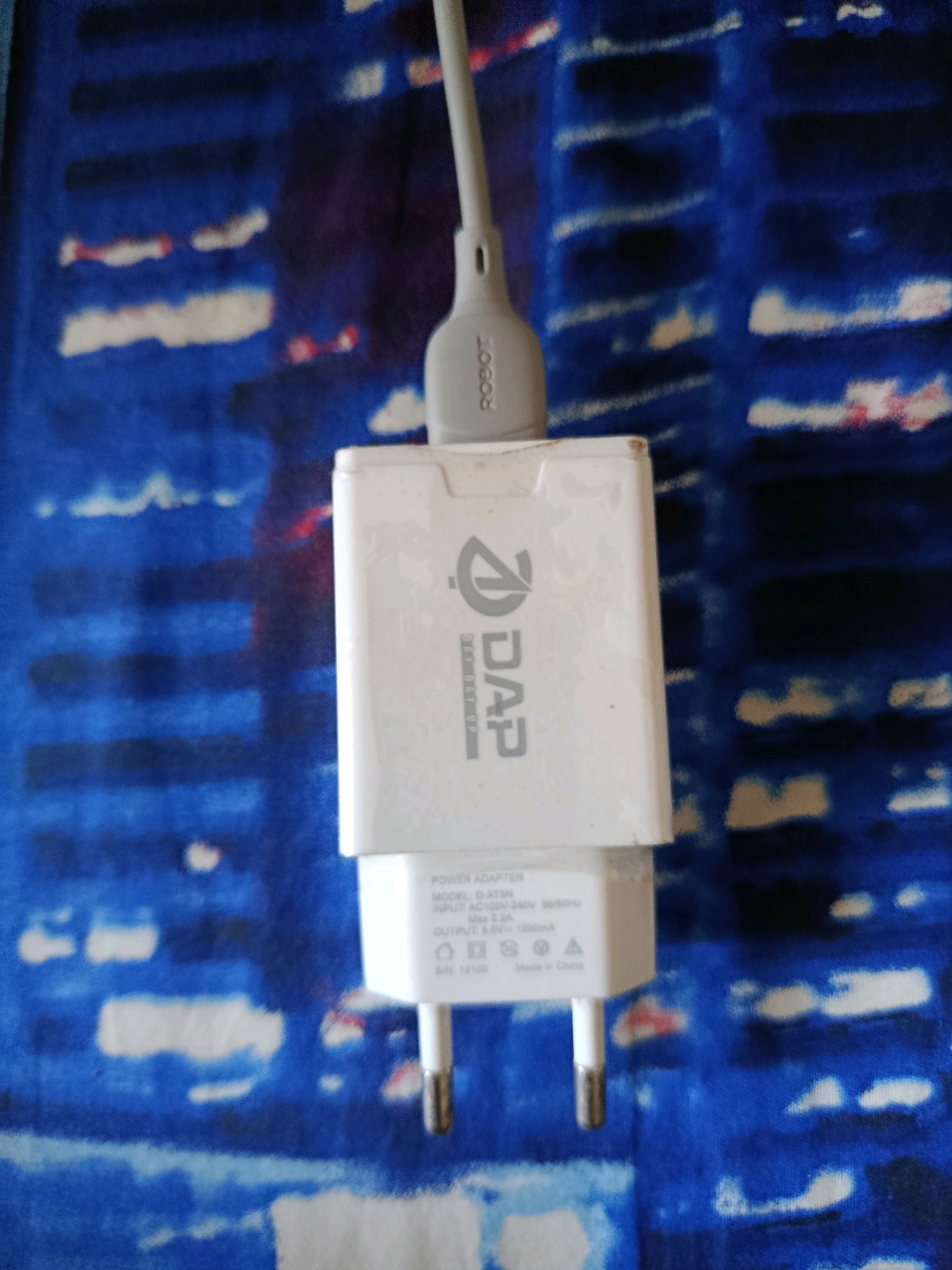 A white wall charger with a gray cable is plugged into an electrical outlet. The charger has the brand name "DAP" printed on it, along with model information and output specifications. The background is a blue and white patterned fabric. The scene appears to be indoors. There are no people visible in the image. The charger is positioned vertically in the frame. The electrical outlet is a standard type with two pins. The cable is a USB cable. The overall impression is one of a functional charging setup.