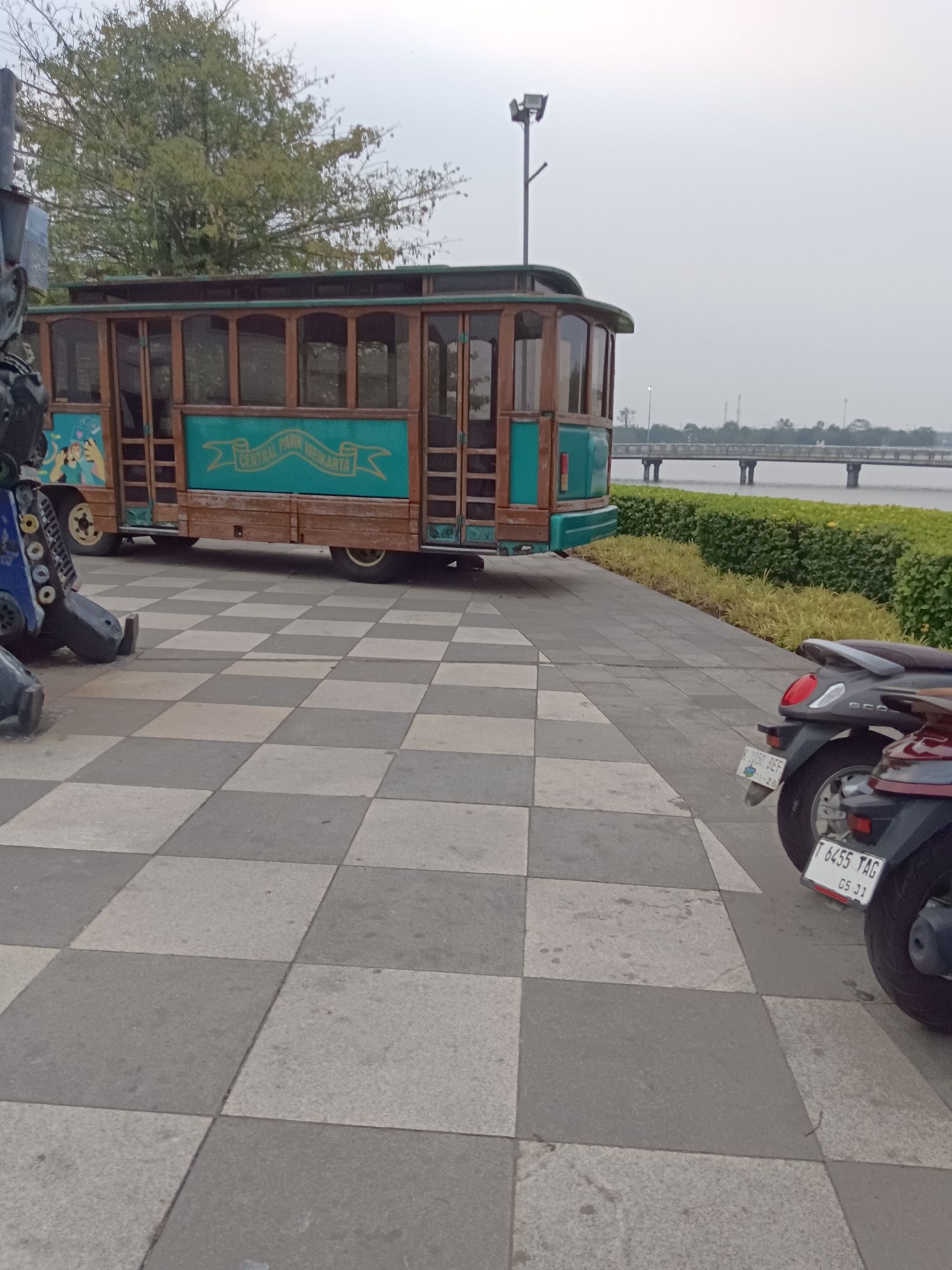The image depicts a scene in the city of Jayamukti, Indonesia, featuring a stationary tram-like structure with a wooden exterior and turquoise accents, positioned on a checkered paved area. The tram has multiple windows and signage that reads "CENTRAL PARK MEIKARTA." To the left of the tram, a portion of a large, dark, metallic robot sculpture is visible. In the foreground, two motorcycles are parked, one dark gray and the other maroon, both with visible license plates. Behind the tram, a body of water is present, with a bridge spanning across it in the distance. Dense green foliage lines the edge of the water, and the sky is overcast.