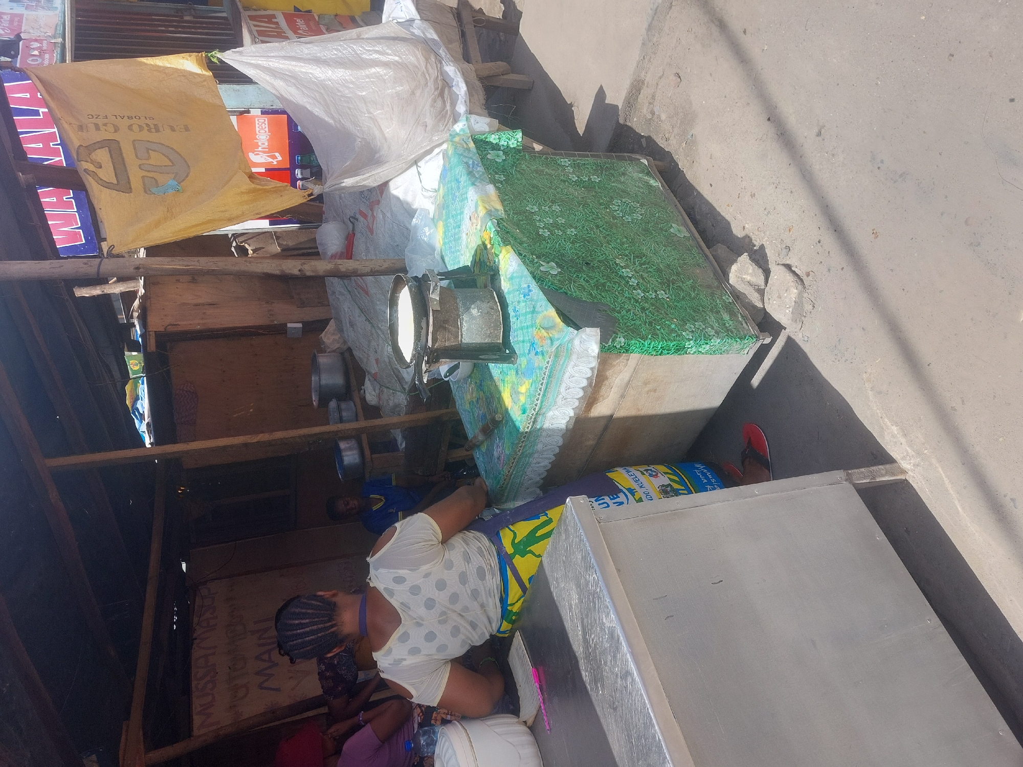 The image depicts an outdoor food stall in Dar es Salaam, Tanzania. A woman wearing a white, polka-dot shirt and colorful skirt is seated behind a table covered with a green and blue flowered tablecloth and a white lace trim. On the table, there is a pot and utensils. Other individuals are present in the background. Above the stall, there is signage including "EURO GUL," "GLOBAL FZC," "MUSSAMASUP," "MAINI," "Airtel," and "Phalo pesa". Other objects visible include cooking pots, bags, and wooden structures. The setting appears to be a busy street.