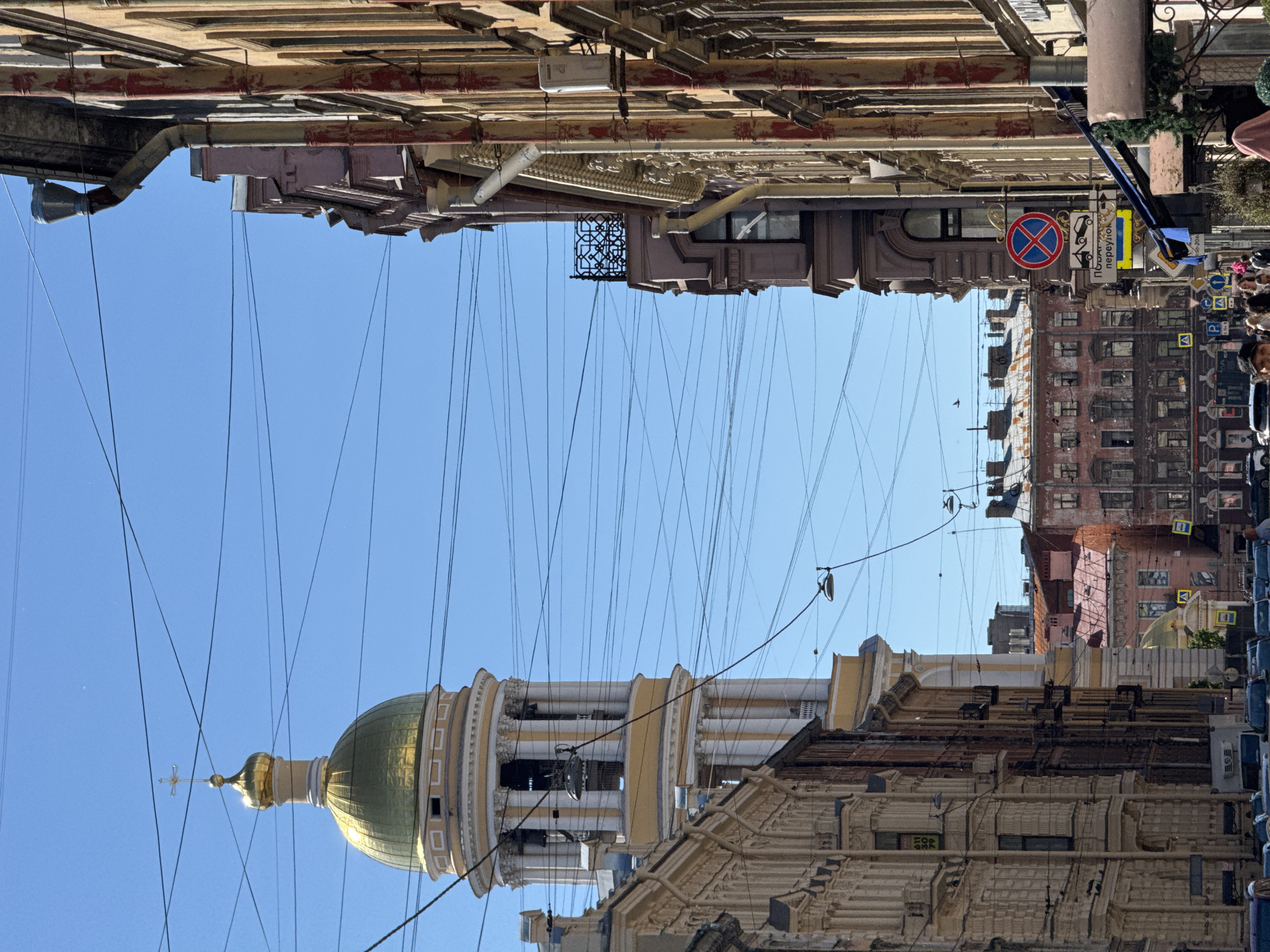 This image depicts a view of a street in Saint Petersburg, Russia, characterized by its ornate architecture and a clear blue sky. The perspective is from a low angle, looking upwards, and the image is rotated 90 degrees counterclockwise.
Prominently featured in the center is the golden dome and belfry of a church or cathedral, topped with a cross. The belfry has a series of open arches and decorative columns. The dome itself is a vibrant gold, reflecting the sunlight. Surrounding the dome are multi-story buildings with intricate facades, typical of historical European city centers. Many of these buildings have ornate cornices, decorative moldings, and classic window designs.
The scene is dominated by a dense network of overhead wires, crisscrossing the sky and obscuring parts of the architecture. These wires suggest the presence of a public transportation system, such as trams or trolleys, or possibly telecommunication lines.
Several street signs are visible on the right side of the image, though the text is largely unreadable due to distance and angle. One sign appears to be a "No Parking" symbol. Another sign, oriented horizontally, shows Cyrillic text.
The lighting suggests it is daytime, likely mid-morning or afternoon, with bright sunlight casting shadows and highlighting the architectural details. The clear sky indicates fair weather.
While no specific activity is explicitly depicted, the presence of street signs and the urban setting imply a bustling city environment. The overall impression is one of historical grandeur and the vibrant, often cable-filled, street life of a European city.