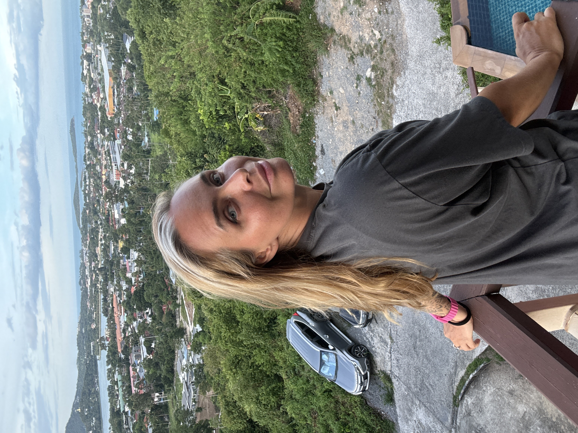 A woman is taking a selfie from a high vantage point, looking down at a coastal town and the ocean. The woman has blonde hair and is wearing a dark grey t-shirt and a pink bracelet on her wrist. Her expression is relaxed and her eyes are looking towards the camera.
The scene shows a densely populated town with a variety of buildings, many with colorful roofs, nestled amongst lush green trees. A body of water, likely the sea, stretches out towards the horizon, with faint clouds in the sky. The ground in the foreground is a mix of gravel and concrete, with a wooden railing. A dark-colored SUV is parked on the gravel area. The overall setting appears to be a tropical location, consistent with Ko Samui, Thailand. The lighting suggests it is daytime, possibly late afternoon.