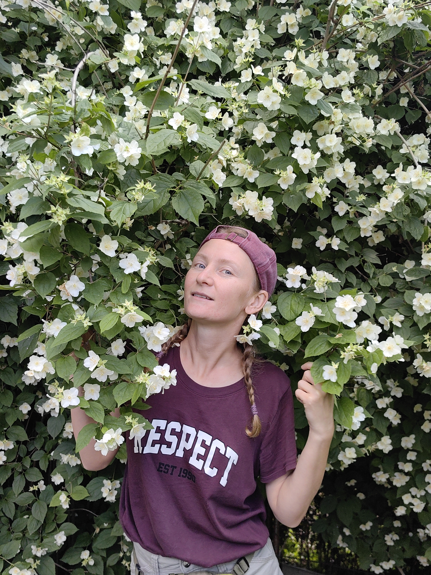 A person with fair skin and braided blonde hair, wearing a maroon t-shirt with the word "RESPECT" printed in white and a maroon cap, stands in front of a dense bush with white flowers and green leaves. The person is looking to the left of the frame with their head tilted slightly. The right hand of the person is holding a branch of the bush. The environment appears to be outdoors, possibly in a park or garden. The image is shot from a medium distance, capturing the person from the chest up and the surrounding foliage. The overall lighting suggests it is daytime. The location in the city of Zuyevo rural settlement, Ukraine, is noted for this image.