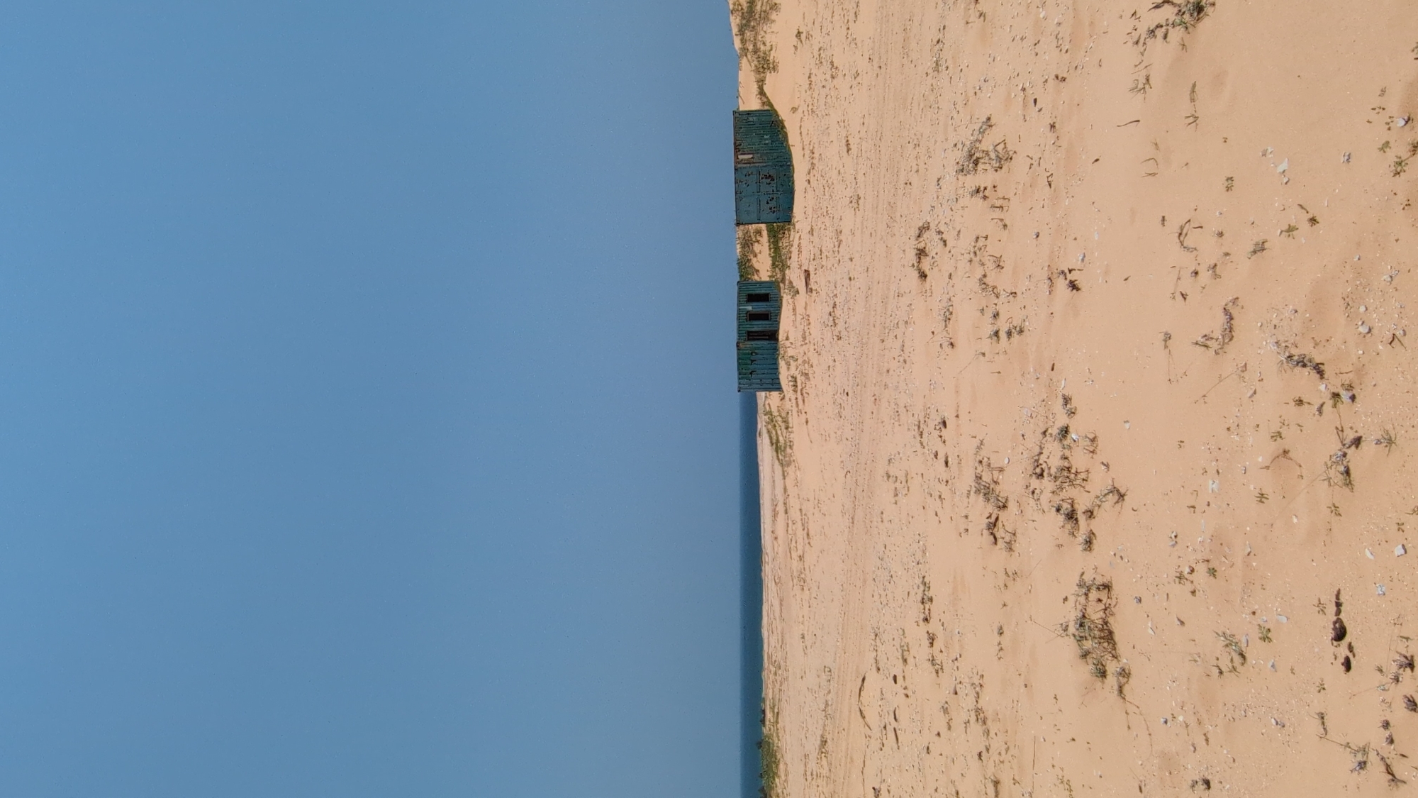 The image depicts a beach scene near Phuoc Dinh, Vietnam. On the right side of the frame, a sandy beach stretches out, marked with sparse vegetation and footprints. Two dilapidated, green metal structures, possibly shipping containers, are positioned on the edge of the beach, near the water. To the left is a clear blue sky. A sliver of the ocean is visible between the beach and the sky. The image is rotated ninety degrees counterclockwise.