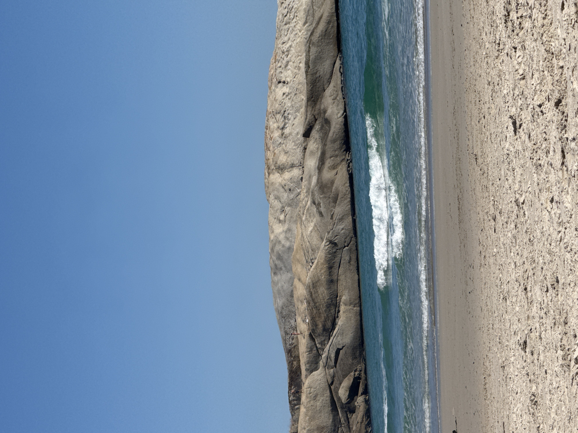 The original image, which is vertically oriented, captures a vibrant coastal landscape under a clear blue sky. When mentally re-oriented to a horizontal view, the scene unfolds from left to right, showcasing a rugged rocky cliff, the expansive ocean, and a sandy beach.
The upper portion of the re-oriented image features a vast, unbroken expanse of light blue sky, indicating bright and clear weather conditions without any clouds.
Dominating the mid-left section is a large, grey, and highly textured rocky cliff face. The rock exhibits clear stratification and weathering, with visible crevices and rough surfaces. A small, red-colored figure, likely a person, is subtly visible on the cliff, suggesting activities such as rock climbing or hiking.
Adjacent to the cliff, in the middle of the frame, lies the ocean. The water exhibits a beautiful transition from clear turquoise near the shore to deeper blues further out. Several white-crested waves are actively breaking and rolling onto the beach, forming lines of white foam along the waterline.
The right side of the re-oriented image presents a wide, light brown sandy beach. The sand appears fine-grained, with scattered shell fragments or small pebbles visible on its surface. A tiny, dark figure, likely a person, can be seen walking near the water's edge on the beach.
The scene conveys a sense of natural grandeur and activity, characteristic of a sunny daytime setting due to the vivid colors and strong natural light. There is no visible text within the image. This picturesque coastal environment is located in Hout Bay, South Africa.