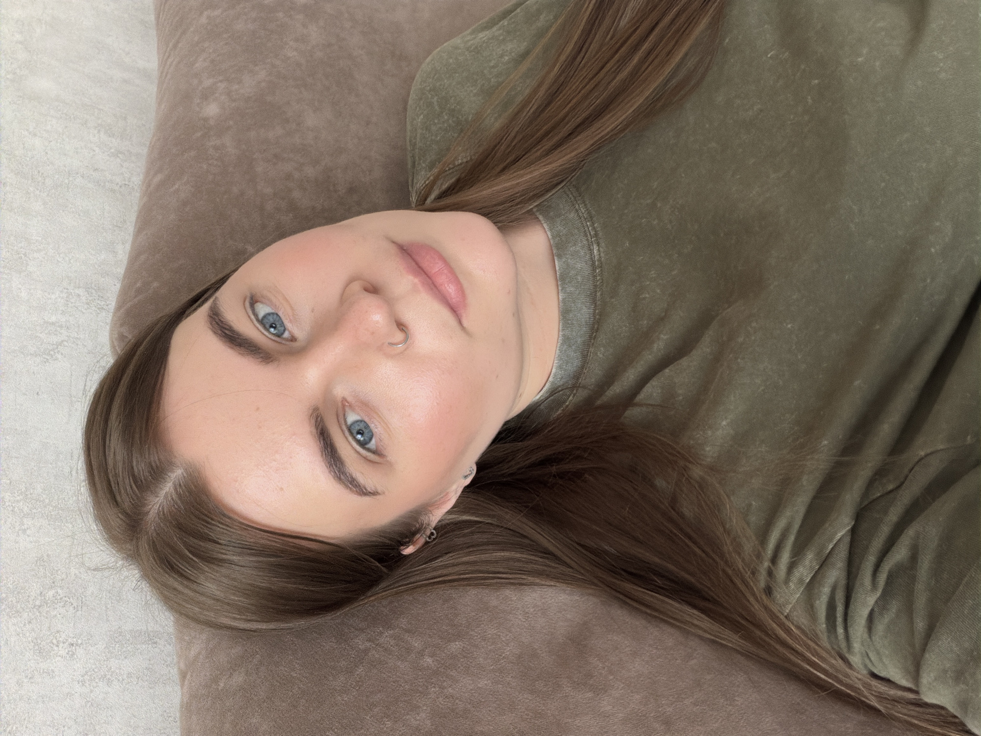 A young woman with light skin, blue eyes, and light brown hair is lying on a brown pillow. She is wearing a green shirt. She has a nose ring and earrings. The background is a light grey surface. The location is unknown, and there is no definitive information to place the scene in Chaikovsky, Russia.