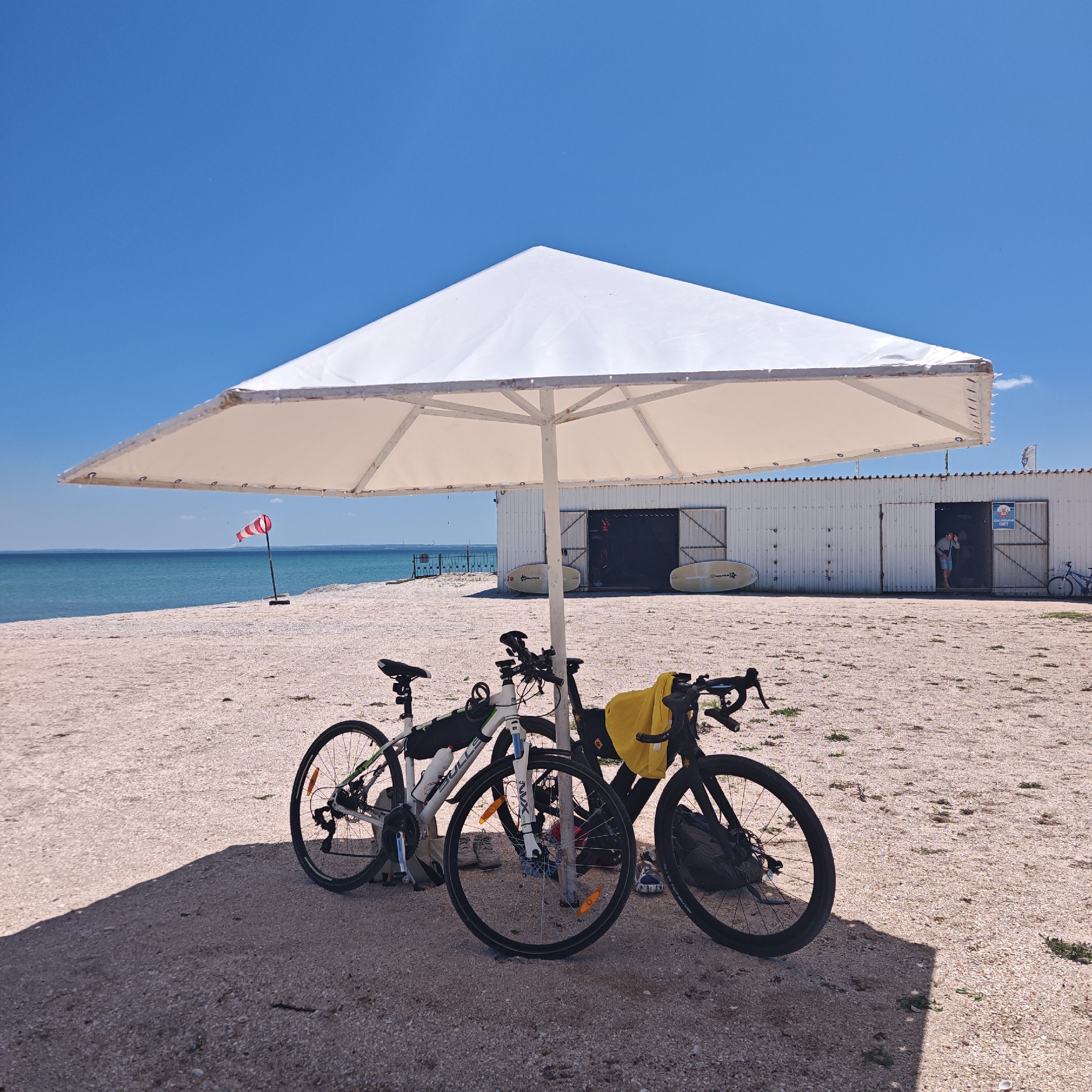 Three bicycles stand beneath a large, white sun umbrella on a beach. The bicycles are of various styles and colors, including white, black, and green, with accessories like water bottles and bags attached. The beach consists of small shells and light-colored pebbles. In the background, the turquoise sea meets a clear blue sky, with a windsock visible near the shoreline. Further back, there is a white, shed-like building with open doors, inside of which are white surfboards. A person is standing in one of the doorways. A sign in Russian, “ПОСТ,” is placed above the door.