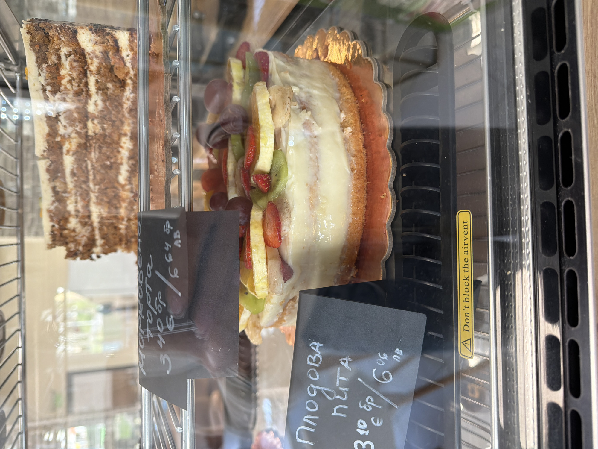 The image captures a display case showcasing two types of cake, presumably in a café or bakery. On the left, a slice of layered cake with brown and white frosting is visible. To the right, a fruit-topped tart or cake is displayed, decorated with grapes, strawberries, kiwi, and pineapple. Price tags are visible in front of each cake; the price tag for the layered cake displays Cyrillic text, which includes the name "MOPKOBE торда," "3:40," and "6.64 ЛВ," with what appears to be "3:40" indicating a price of 3 Euros 40 stotinki, and 6.64 levs. The price tag for the fruit tart reads "Плодова NUTA," "3:10 €", and "6:06 ЛВ," indicating "Fruit Pita", a price of 3 Euros 10 stotinki, and 6.06 levs.
The display case features a warning sticker that reads "Don't block the airvent".
The scene likely takes place in a city where both Euro and the Bulgarian lev are accepted as currency. Based on the Cyrillic text, it is probable that the location is within Bulgaria, such as the city of Gabrovo.