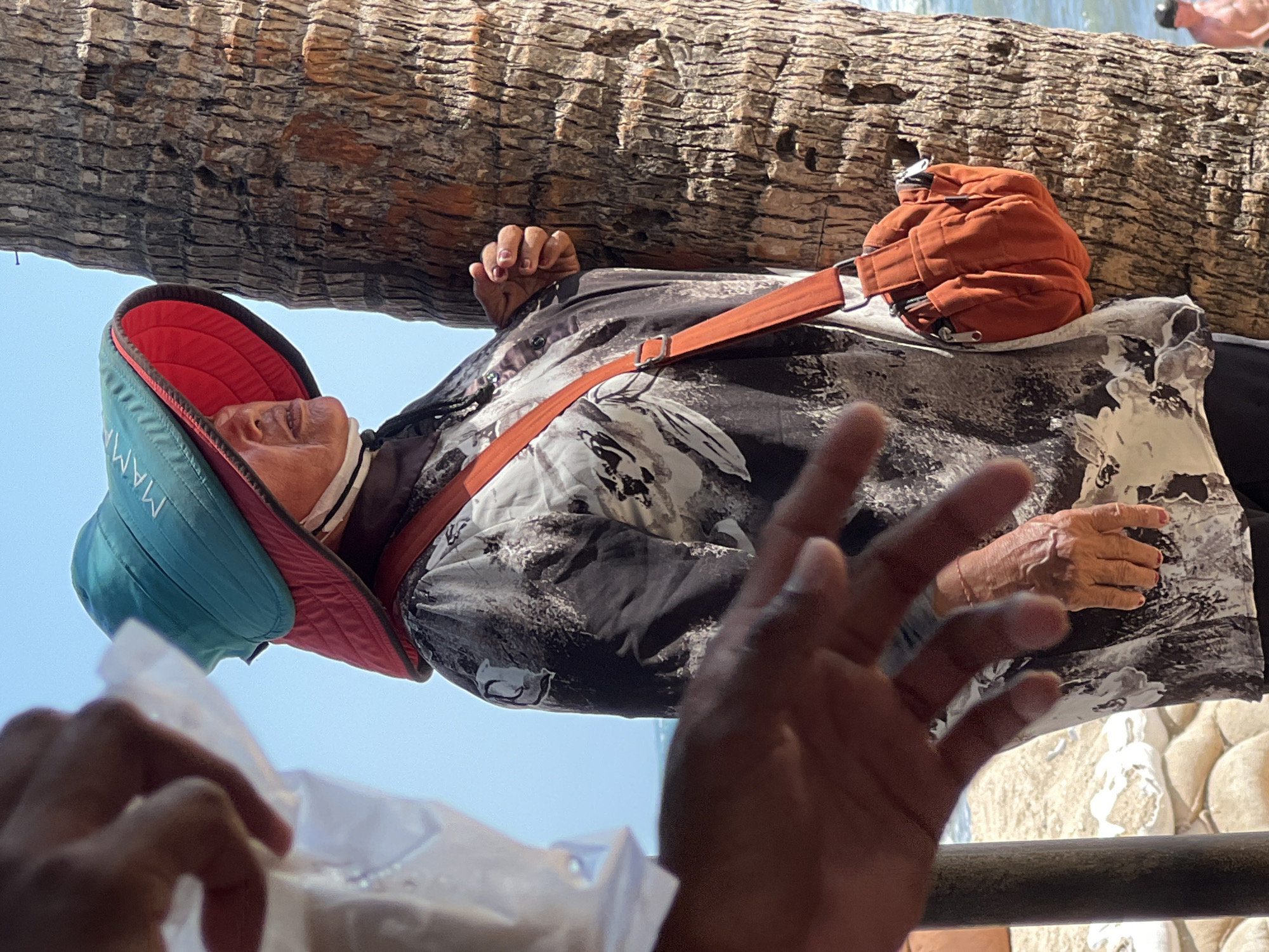 The image depicts a woman standing next to a palm tree with the ocean visible in the background. She is wearing a large, wide-brimmed hat that is teal on top and red underneath, with white text that reads "MAMA." Her face is partially obscured by the hat, but she appears to be looking to the right. She is also wearing a dark, patterned shirt and an orange crossbody bag. In the foreground, out of focus, are two hands. The sandbags in the background suggest a beach setting. This scene is consistent with locations in Kuta, Indonesia, known for its beaches and coastal activities.