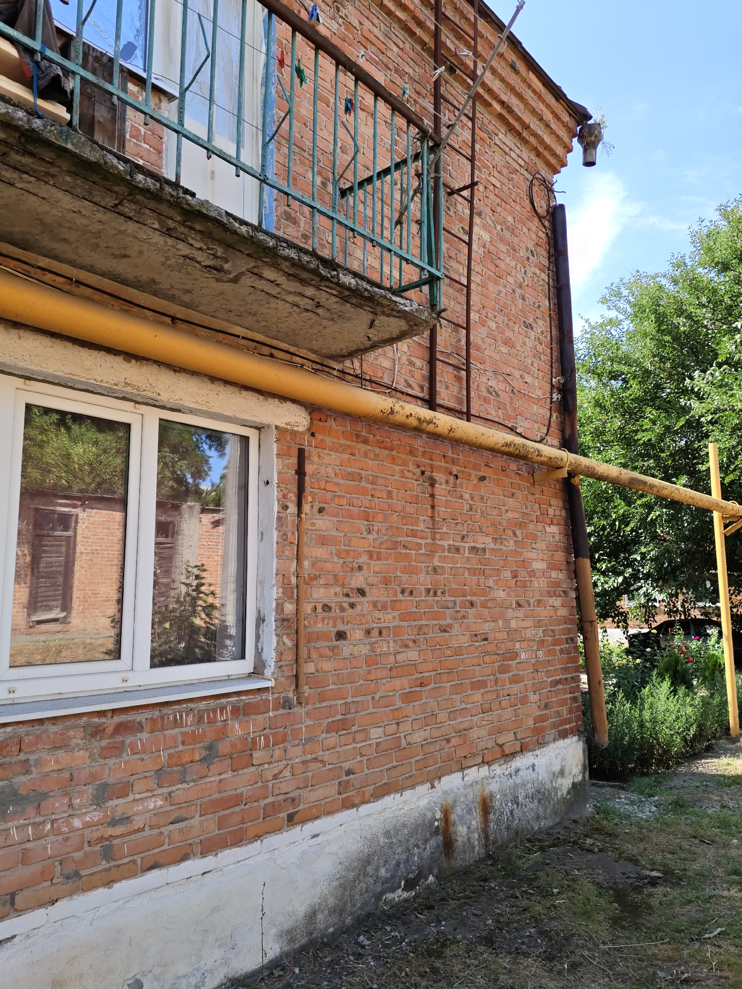 The image depicts the exterior of a brick building with a prominent yellow pipe running horizontally along its side. A window with white frames is visible on the lower left. Above the window, a concrete balcony with a green metal railing is present. The building's brickwork is varied in color and shows signs of age. A ladder is attached to the side of the building, leading up towards the roofline. A drainpipe runs vertically along the brick wall. The scene is set outdoors with greenery and trees visible in the background, suggesting a residential or semi-rural area. The sky is clear and blue with some clouds. The ground appears to be a mix of grass and dirt. The overall impression is of an older, functional building in need of maintenance. There are no people or animals visible in the image. The location is identified as Pukhlyakovskiy, Russia.