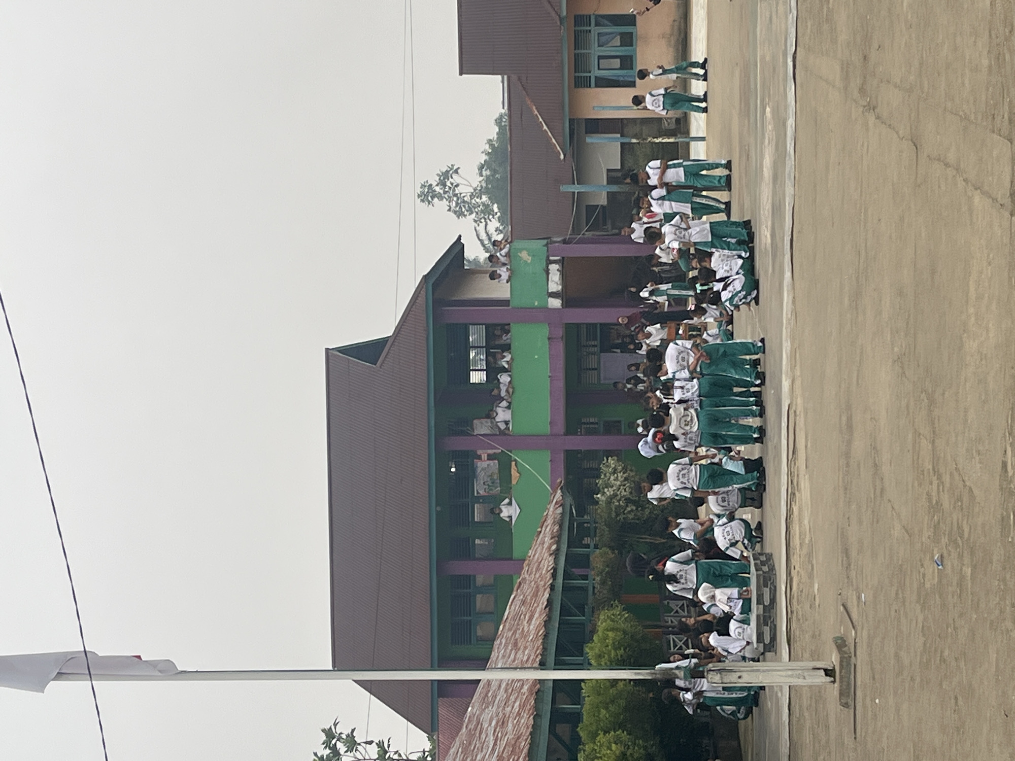 This image captures a scene at a school in Meliau Hulu, Indonesia, possibly during an outdoor activity or assembly. The focal point is a group of children, predominantly wearing green and white uniforms, gathered in a courtyard. Several children are seated on the ground or on a raised platform, while others stand in groups. Some children are looking towards the camera, while others are engaged in conversation or activity amongst themselves.
Behind the children, a two-story building with a dark brown roof and green and purple accents is visible. This building appears to be part of the school's infrastructure, with balconies or open-air spaces where more children and adults are observing the scene below. Another smaller building with a similar roof is located to the left of the main building, and a more distant building with a light brown exterior and a blue window is visible on the right. A white flagpole stands prominently on the left side of the image. The overall environment suggests a school campus with a somewhat overcast sky, indicating it might be morning or afternoon. The ground is paved with concrete, showing some cracks and a general dusty appearance. The scene appears to be calm, with children interacting in a relaxed manner.