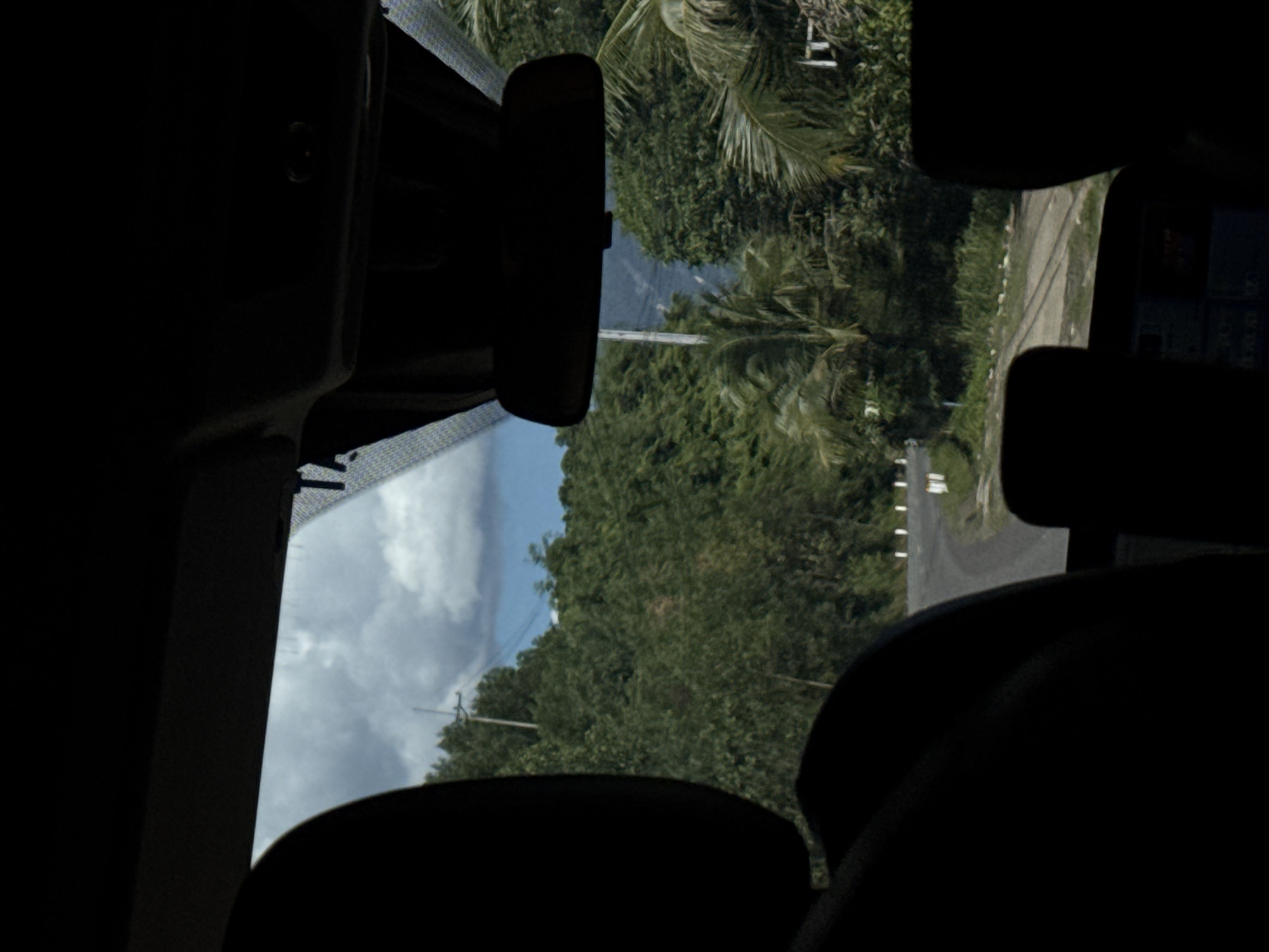 A view from inside a vehicle looking out through the windshield at a rural road winding through a lush, tropical landscape. The foreground is dominated by the dark silhouettes of car seats and the interior of the vehicle. The windshield reveals a cloudy sky with patches of blue. Beyond the windshield, a paved road curves to the left, flanked by dense green foliage. Tall trees, including palm trees, create a verdant canopy. A single utility pole with wires is visible in the distance. The scene appears to be in a rural area of Nam Khánh Vĩnh, Vietnam, characterized by its abundant vegetation and the winding road suggesting a mountainous or hilly terrain.