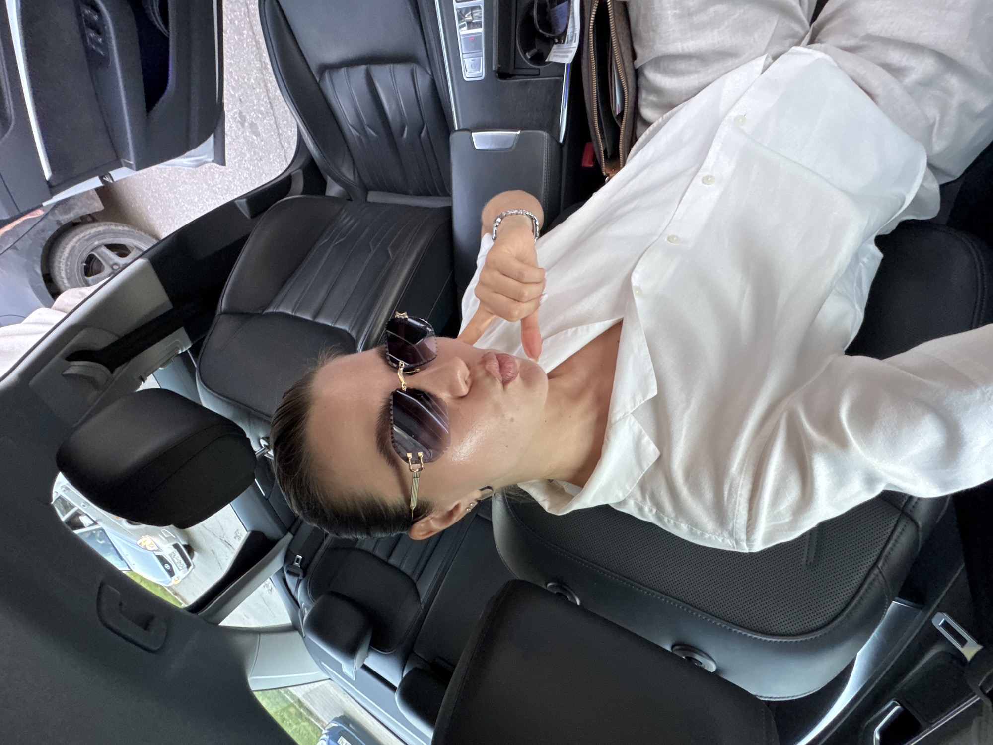 A woman is sitting in the back seat of a car and taking a selfie. She is wearing a white button-down shirt and sunglasses. She is puckering her lips and pointing to her chin with her right hand. The car interior is visible, with black leather seats and a center console. Outside the car, another car is visible, as well as some greenery. The scene appears to be in a city, but the specific location is unknown.