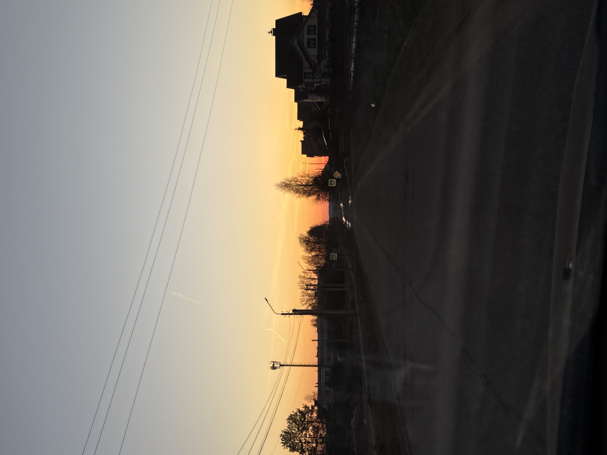 The image depicts a street in Прикамский, Russia, at sunset. A road stretches into the distance, flanked by buildings on one side and trees and utility poles on the other. A street sign is visible near a cluster of trees. Power lines run across the sky. The sky is transitioning from light blue to orange. The photo is taken from inside a vehicle, with reflections visible on the window.