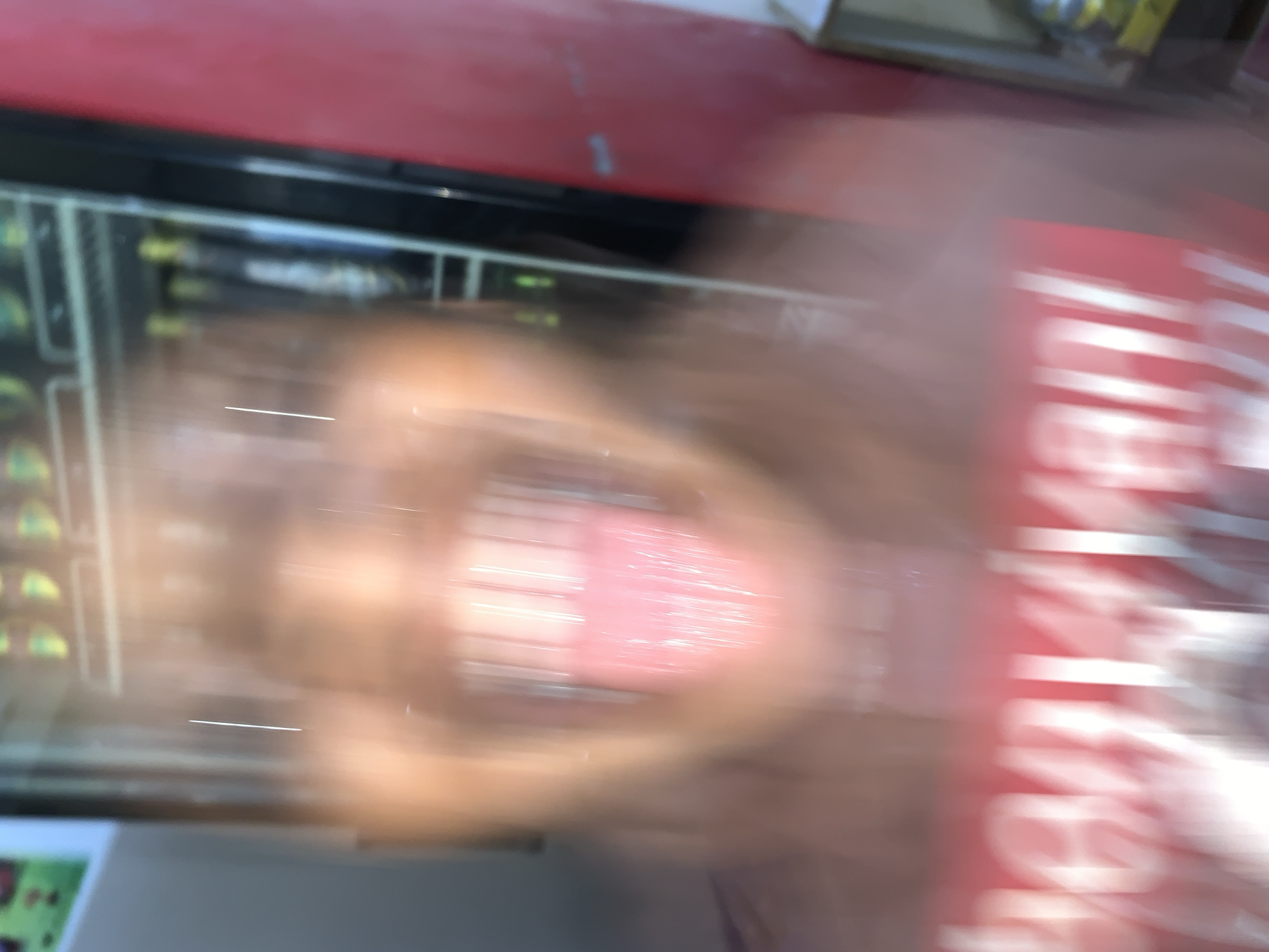 The image depicts a heavily motion-blurred, close-up view of an individual within an indoor commercial setting in New Stadium, Nigeria. The central subject is a person, whose facial features are largely indistinct due to extreme blur, but a warm skin tone is observable. The person's mouth is wide open, displaying rows of blurred, light-colored teeth and a distinctly prominent, extended tongue. The tongue appears vibrant pink-red, characterized by pronounced light streaks indicative of moisture and motion. Below the person's chin, a red garment with large, blurred white lettering is visible. In the background, a large, commercial refrigerated display unit or vending machine is partially visible, containing multiple shelves stocked with indistinct items, possibly beverages, with some displaying yellow and dark coloration. A vertical light streak, likely a reflection or internal light, bisects the background. To the far right, a solid red panel or wall section is present, exhibiting minor surface streaks. The upper left corner shows faint, blurred green and red shapes, possibly from a poster or decal.
