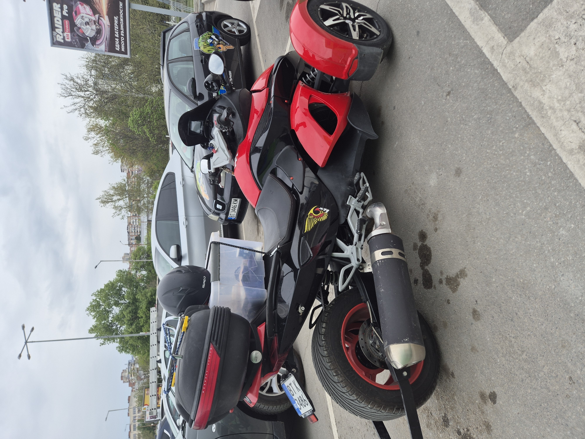 An outdoor scene in Veliko Tarnovo, Bulgaria, features a red and black three-wheeled vehicle, likely a trike or scooter, parked on asphalt under an overcast sky. The vehicle is equipped with a large black and silver exhaust, a black helmet (text "ISPIDO") resting on the back, and black side bags (partially visible text "MORRIFT SYSTEM"). A clear plastic storage box is strapped onto the rear, and a yellow and red bird emblem is visible on its side panel. The rear wheel has a red rim, and a red fender guards one of the front wheels. The license plate reads "BT 3446 KM", with "BT" indicating Veliko Tarnovo.
Behind the trike, a black car with a green and yellow graphic and a "ТЪРНОВО" (Tarnovo) sticker is parked, and a silver car is partially visible. In the background, green trees, distant buildings, and utility poles are present. A billboard prominently displays "RAIDER R20 System Pro" and the Bulgarian text "ЕДНА БАТЕРИЯ, МНОГО ВЪЗМОЖНОСТИ!" (ONE BATTERY, MANY POSSIBILITIES!). The setting appears to be a parking lot or street, devoid of people.