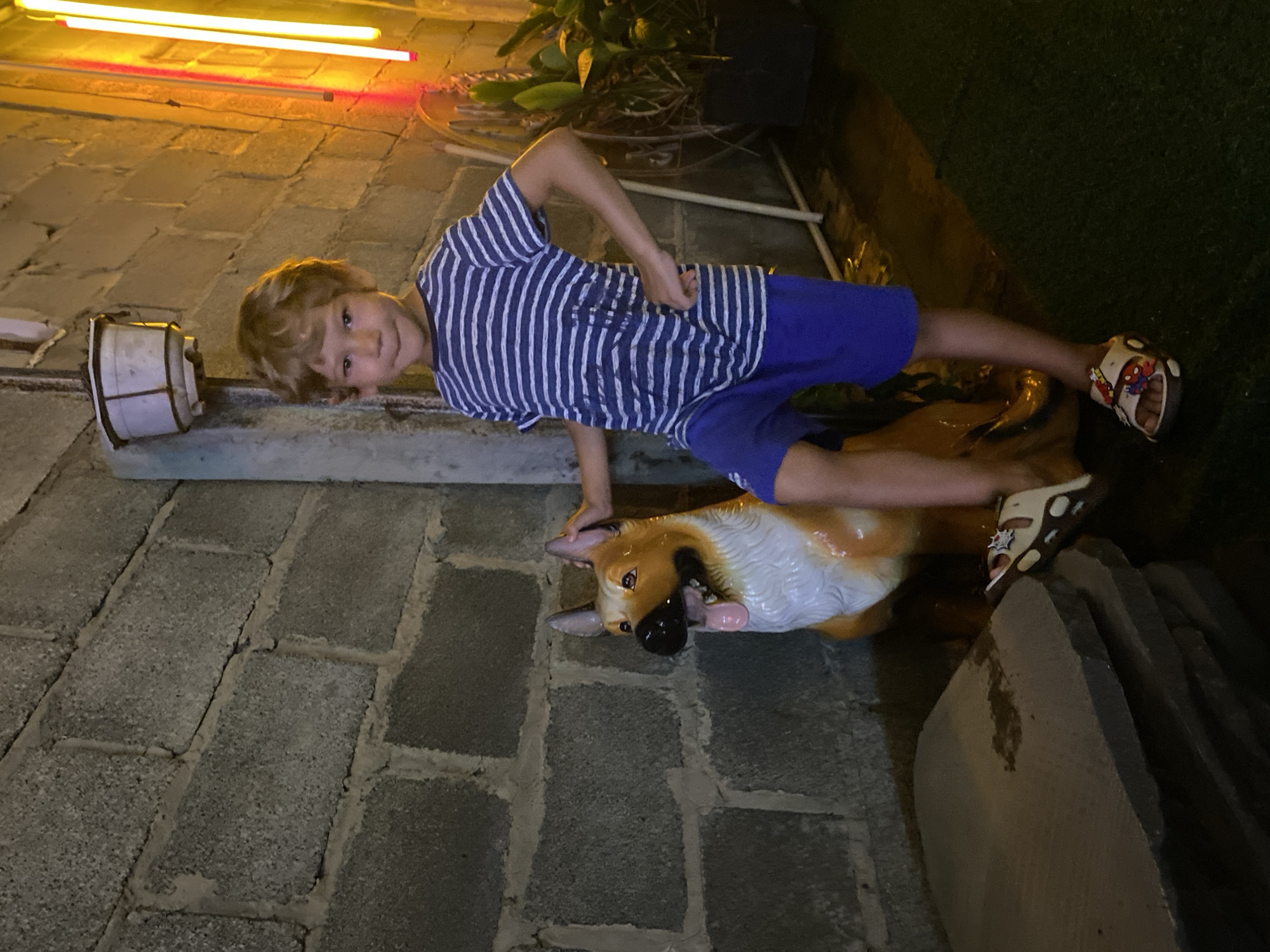 The image depicts a young boy standing next to a statue of a German Shepherd dog. The boy is wearing a blue and white striped t-shirt and blue shorts, and has his left foot propped on the dog statue. The statue is lifelike, with its mouth open and tongue hanging out. The background consists of a grey cinder block wall and some greenery. There are also some bright yellow neon lights visible on the right side of the image, suggesting an urban or commercial setting. The ground appears to be covered in artificial turf in some areas. The scene is dimly lit, with artificial lighting contributing to the overall atmosphere. It is possible this location is a park, a public space, or an outdoor seating area of a restaurant or cafe in Mũi Né, Vietnam, given the presence of lush greenery and the general ambiance.
