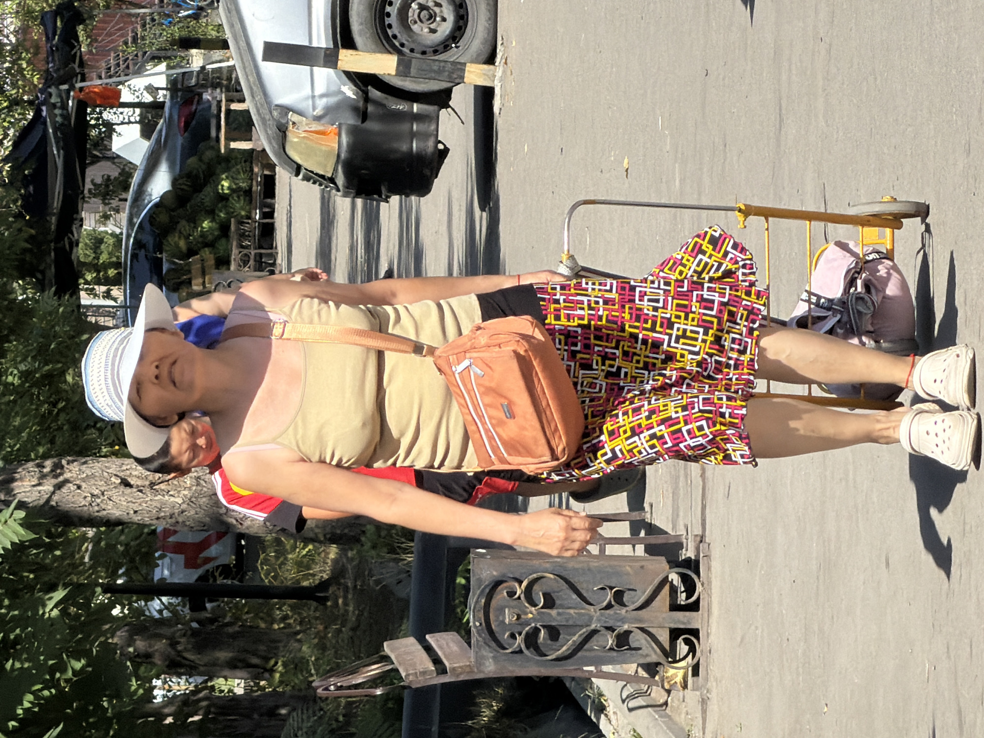 A medium shot captures a woman walking with a small wheeled cart. She is wearing a white and blue striped sun hat, a tan camisole, and a colorful, geometric-patterned skirt. A coral-colored cross-body bag is slung over her shoulder. Her feet are shod in light-colored clogs. She is pushing a yellow wheeled cart that carries pink and gray bags. Behind her, a young person wearing a red and black shirt is partially visible. The scene appears to be outdoors, with a paved walkway and trees in the background. To the right, a dark-colored van is parked. A decorative metal trash receptacle and a wooden bench are positioned to the left of the woman. The lighting suggests it is daytime, with shadows cast on the ground.