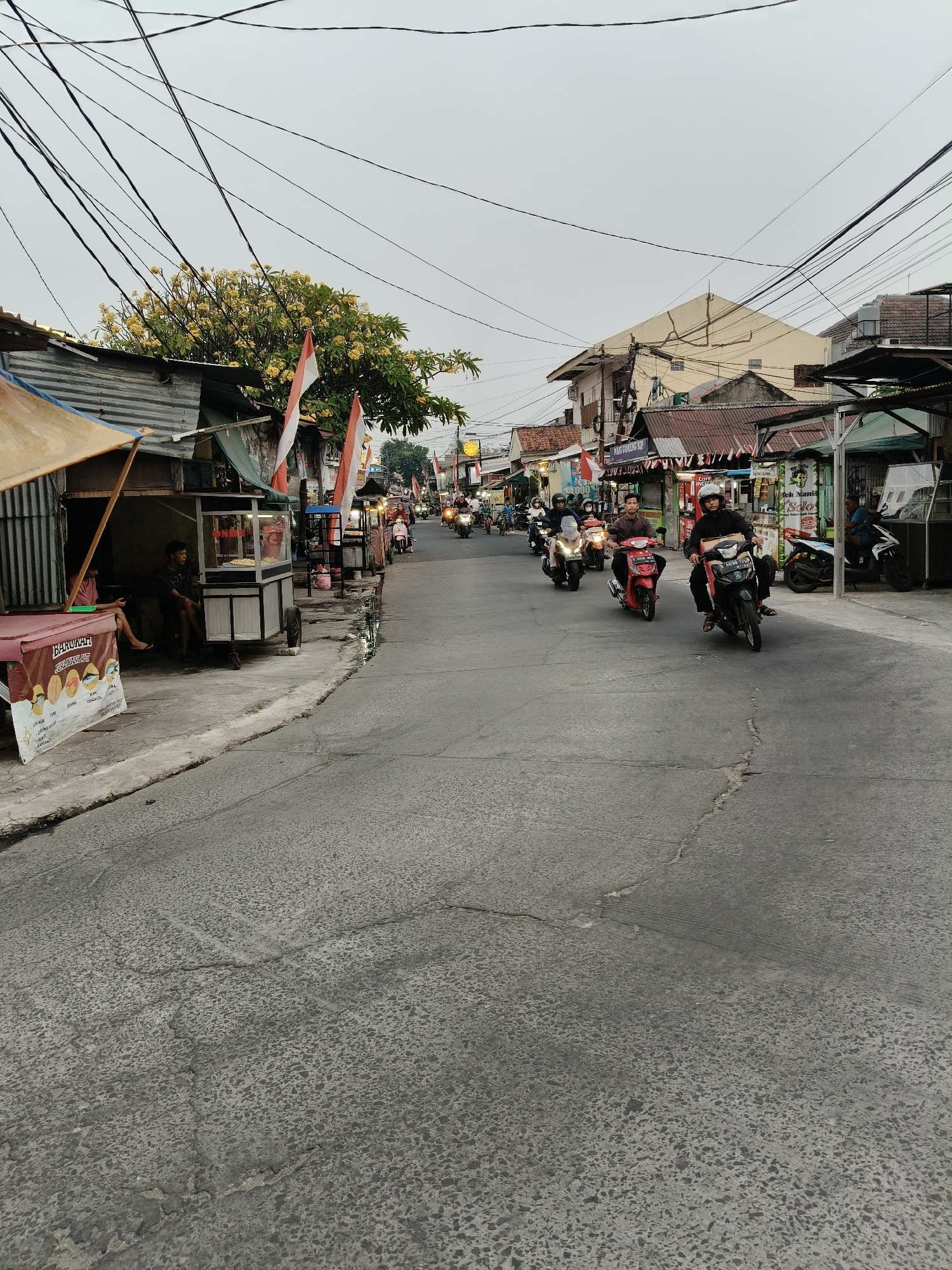 The image depicts a street scene in a residential or commercial area, likely in South Tangerang, Indonesia, characterized by its bustling atmosphere and evident local culture. The sky is overcast, suggesting either early morning or late afternoon light. The street is paved with asphalt and shows signs of wear and tear, with visible cracks and patches.
On the left side of the street, there are small shops or stalls with corrugated metal roofs and awnings. One stall features a red banner with white text and images of food items like fish, and appears to be selling some form of prepared food. A few individuals are seated or standing near these stalls. There are also small carts or kiosks, one of which appears to be selling snacks displayed in a glass case. Indonesian flags are prominently displayed on some of the buildings, hinting at a national holiday or celebration.
The right side of the street is lined with more buildings, also appearing to be shops or houses, with various architectural styles and roof types. Overhead, a complex network of black electrical wires crisscrosses the sky, a common sight in many urban and suburban areas in Indonesia.
The street is active with motorcycle traffic. Several people are riding motorcycles, some with helmets. The motorcycles vary in color and model. The general impression is of a typical street in a populated area of Indonesia, with a mix of residential and commercial establishments, a lively street life, and the ubiquitous presence of motorcycles as a primary mode of transportation.