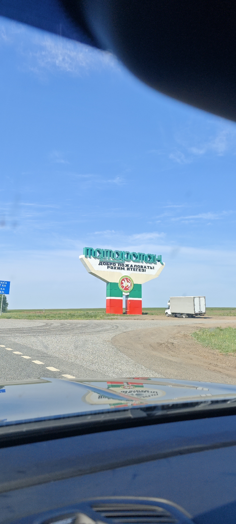 The image depicts a roadside scene in Камаево, Russia. A large sign with the words "TATARSTAN" in Cyrillic script and "WELCOME" in Russian and Tatar stands beside the road. The sign features the flag of Tatarstan. A small white truck is parked nearby. The view is from inside a vehicle, with the dashboard visible in the foreground. The sky is blue and clear. The ground is flat and grassy.