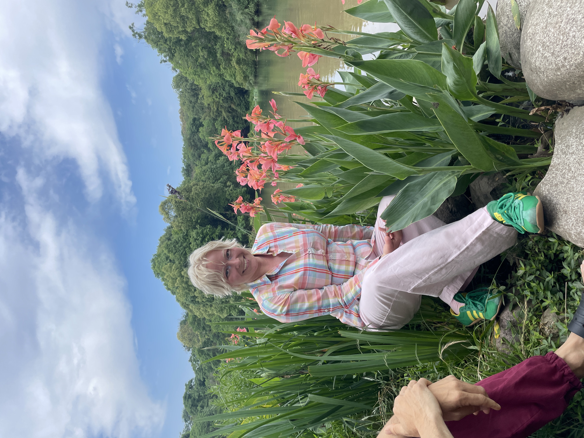 The image shows a woman seated amidst flowers and greenery, likely near a body of water given the glimpse of a lake or pond in the background. She is wearing a multi-colored plaid shirt and light pink pants, with green sneakers on her feet. The flowers are tall and orange/pink in color. The setting appears to be a park or garden. Part of another person is visible in the lower right corner of the image. The sky is visible with a mix of blue and clouds. The location is Chenghua District, China.